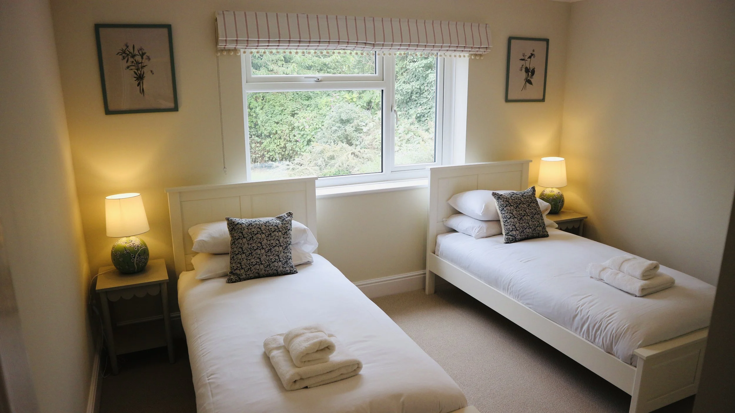 A cosy bedroom with two single beds, each with white bedding, pillows, and folded towels. There are two bedside tables with lamps, framed botanical art on the walls, and a large window overlooking the garden. Blackbird Cottage, recently refurbished.