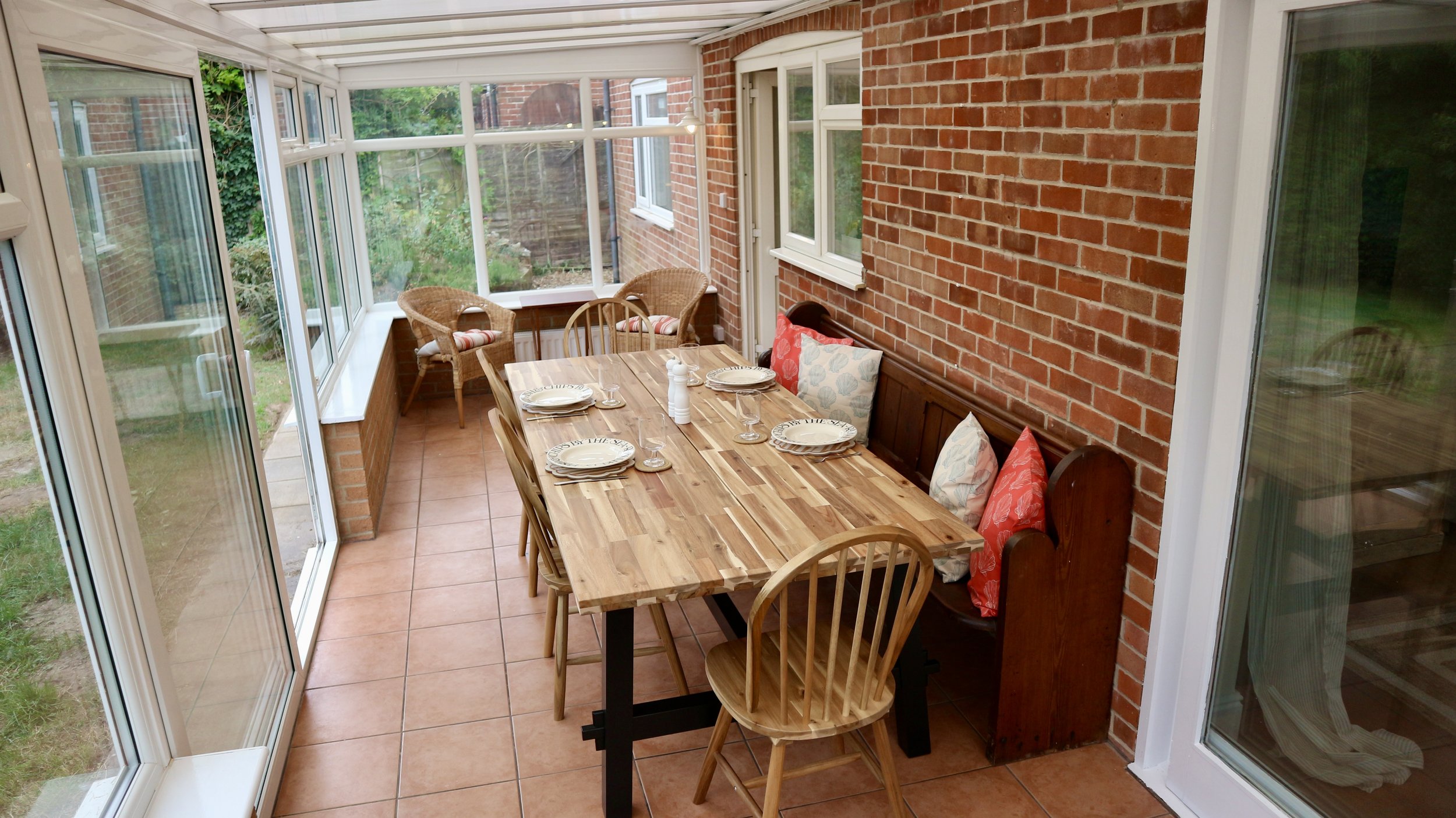 A sunroom with a wooden dining table set with plates, glasses, and a salt shaker, surrounded by wooden and wicker chairs, with brick walls and large windows showing a garden outside.