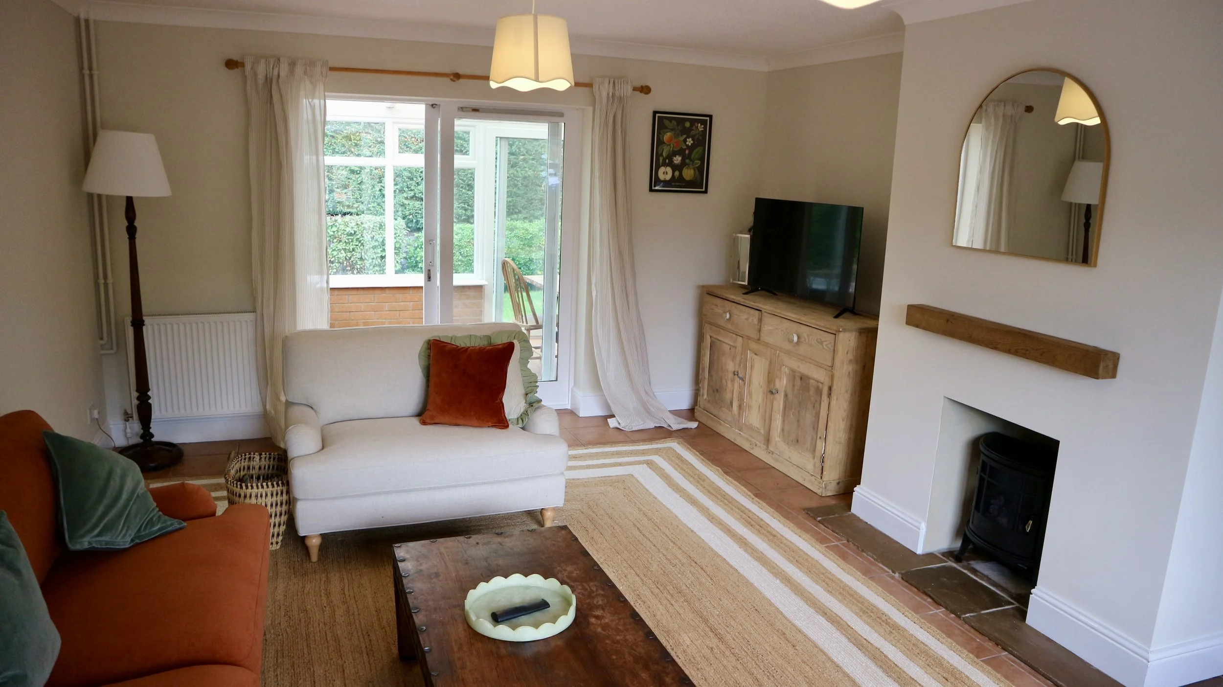 Living room with cream and orange sofas, green and orange pillows, a wooden coffee table with remote, striped rug, sliding glass door leading to a porch with a wooden chair, a TV on a wooden cabinet, a mirror above a fireplace, a floor lamp, and curt
