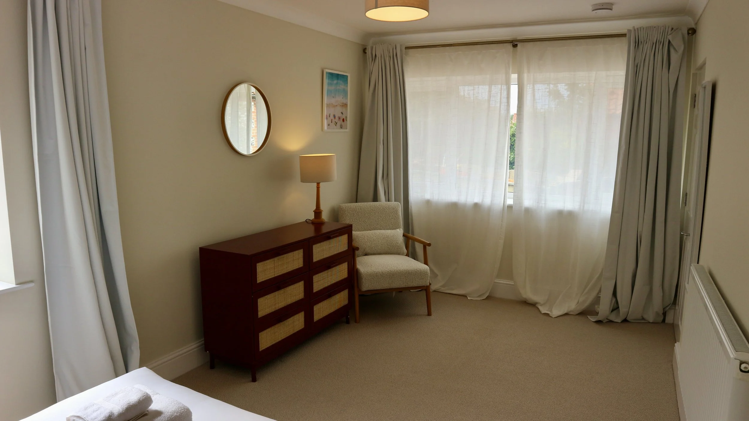 A cosy bedroom corner with beige walls, a window with sheer curtains, a mid-century modern armchair, a red rattan chest of drawers, a table lamp, a round mirror, and a framed seaside artwork. Blackbird Cottage, recently refurbished.