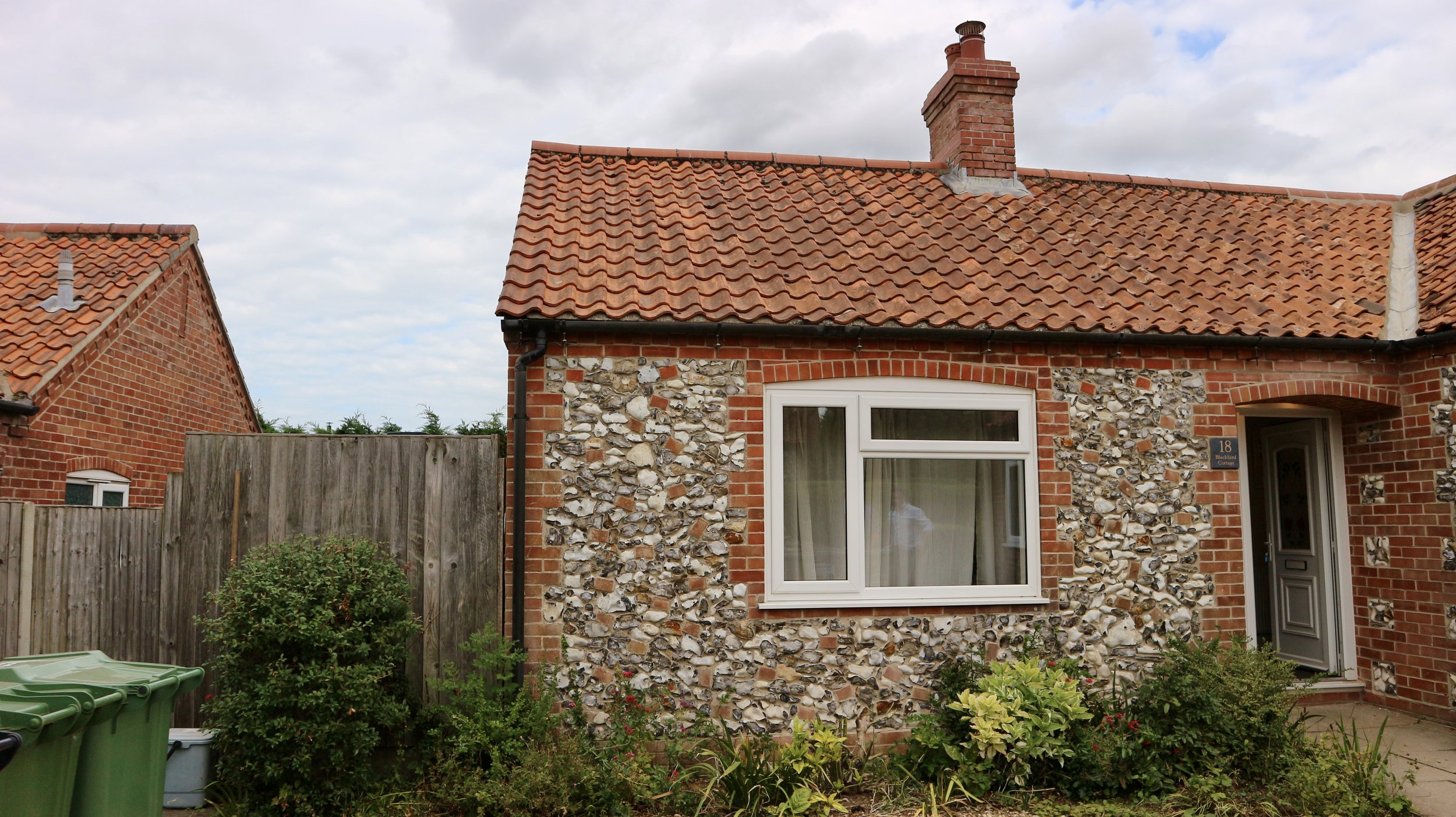 Front of a Blackbird Cottage, recently refurbished, with a tiled roof, a large front window, a front door, and a small garden with bushes and flowers. A wooden fence separates its side yard from neighbouring houses.