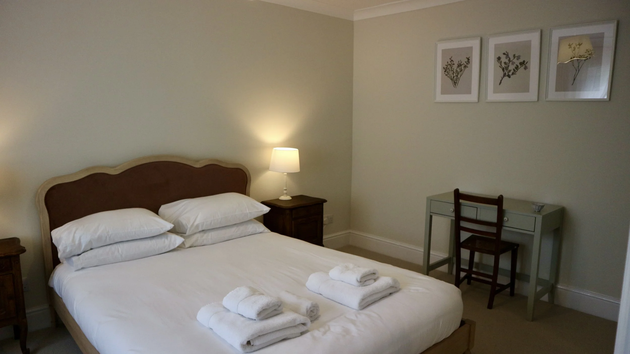 The Master Bedroom with a bed, pillows, and towels, a nightstand with a lamp, and a small desk with a chair, three framed botanical prints on the wall. Blackbird Cottage, recently refurbished.