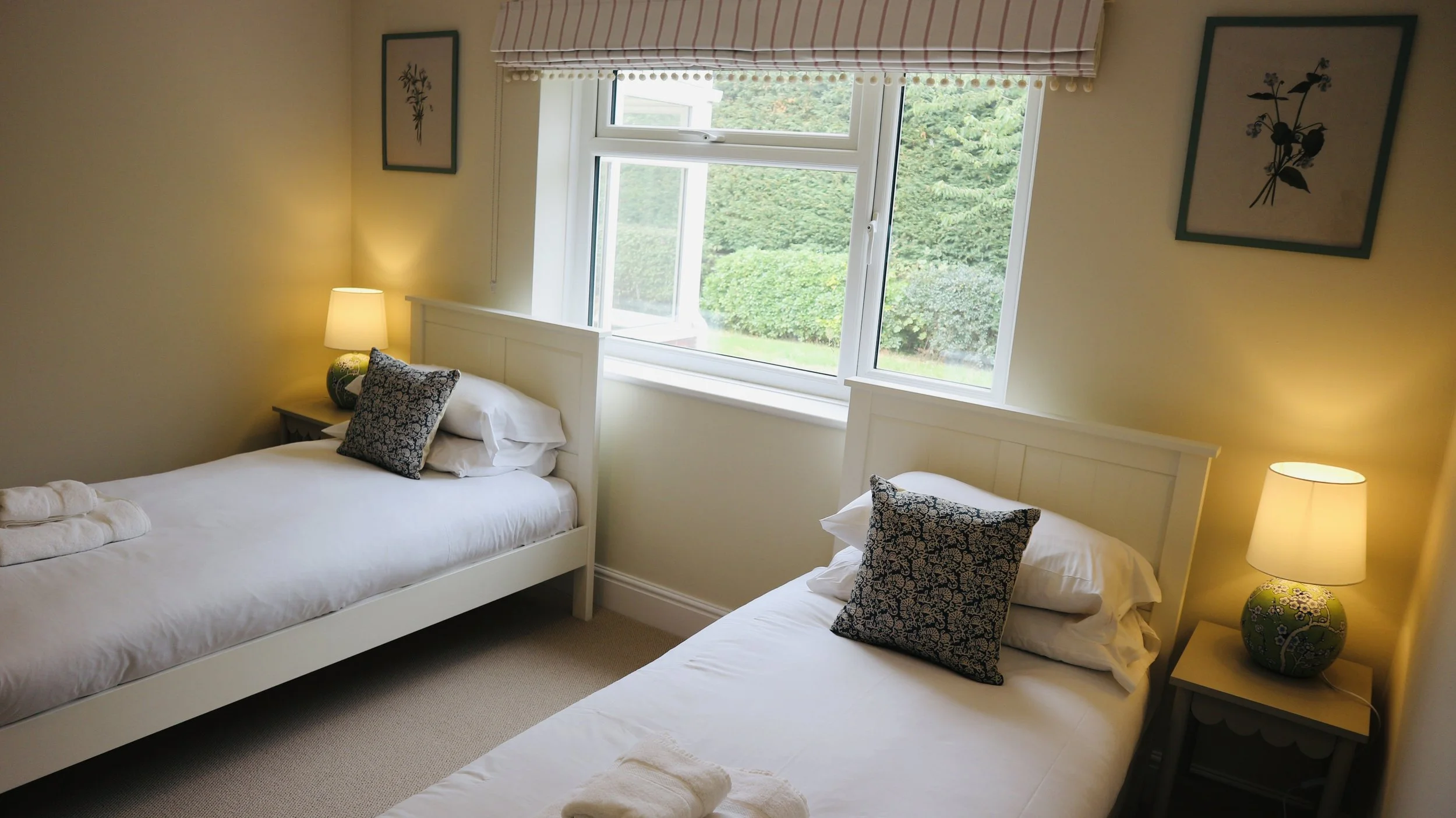 Two single beds in a bedroom with white wooden headboards, dark patterned pillows, bedside lamps, a window with a view of green trees, and framed botanical pictures on the wall. Blackbird Cottage, recently refurbished.