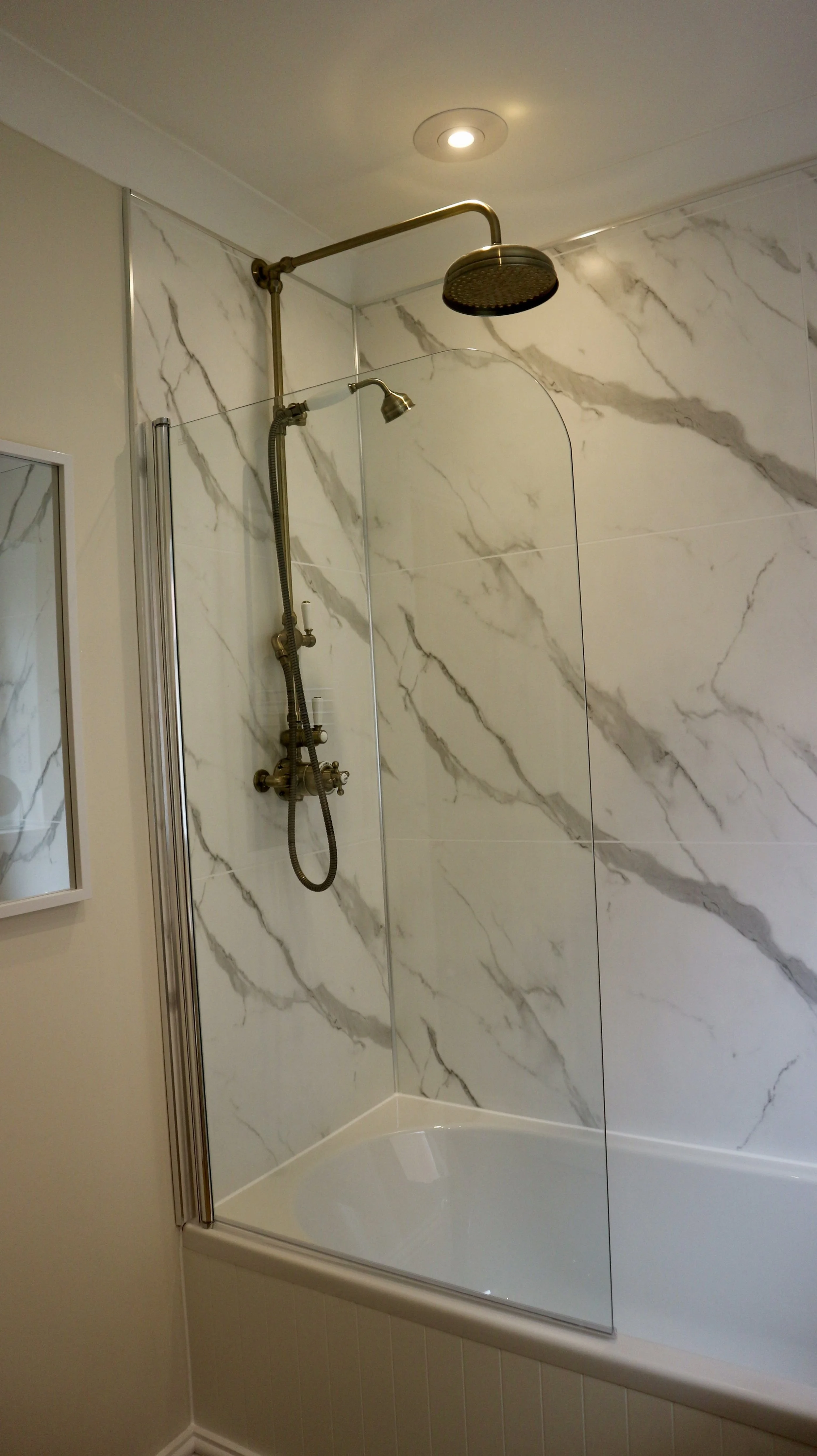 Bathroom shower with marble tile walls, a brass showerhead, handheld shower wand, and a clear safety glass shower screen. Family bathroom at Blackbird Cottage, recently refurbished.