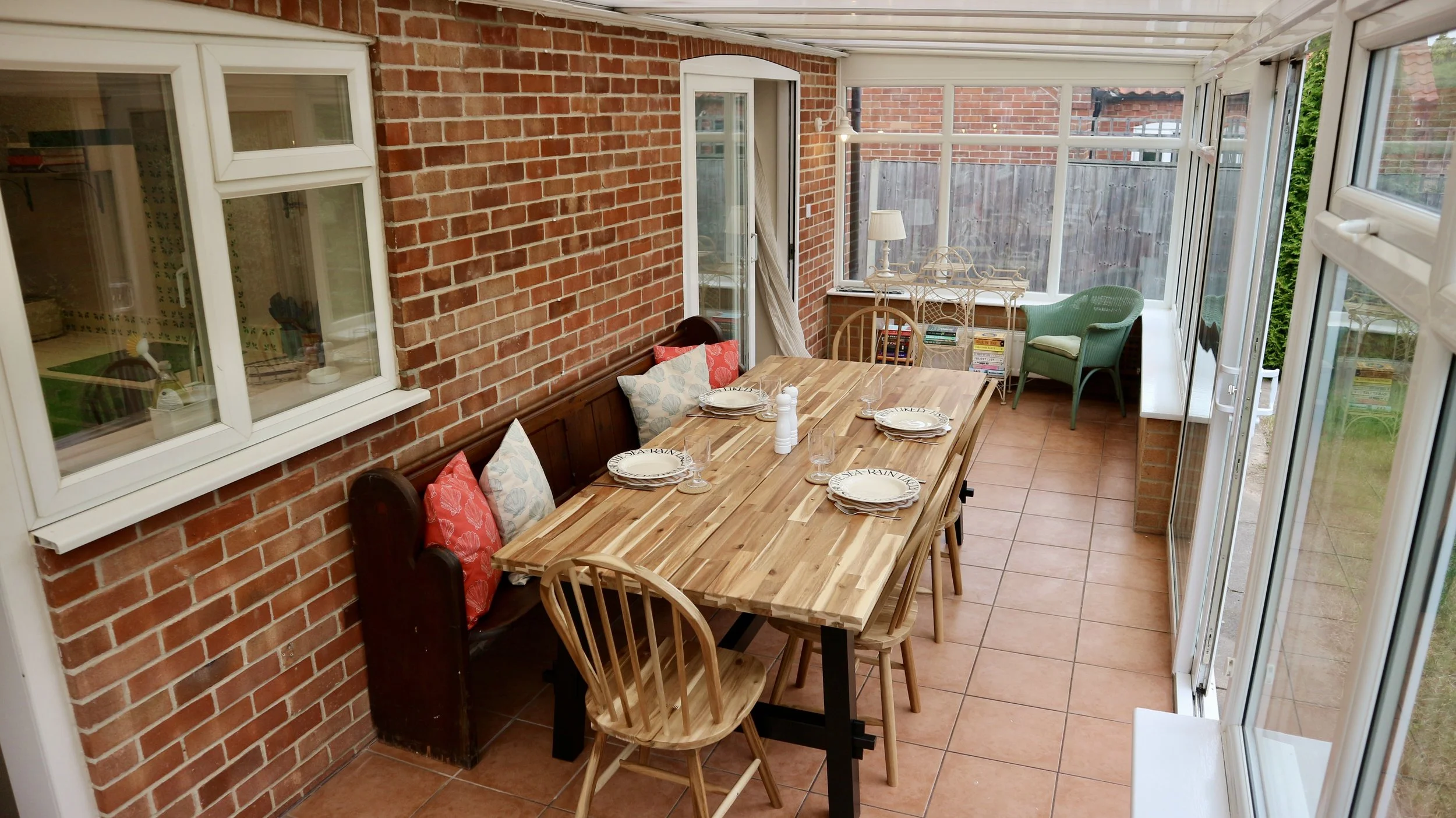 Conservatory with a wooden dining table set for four, surrounded by a brick wall and white-framed windows, with chairs, cushions, a side table, and decorative items.