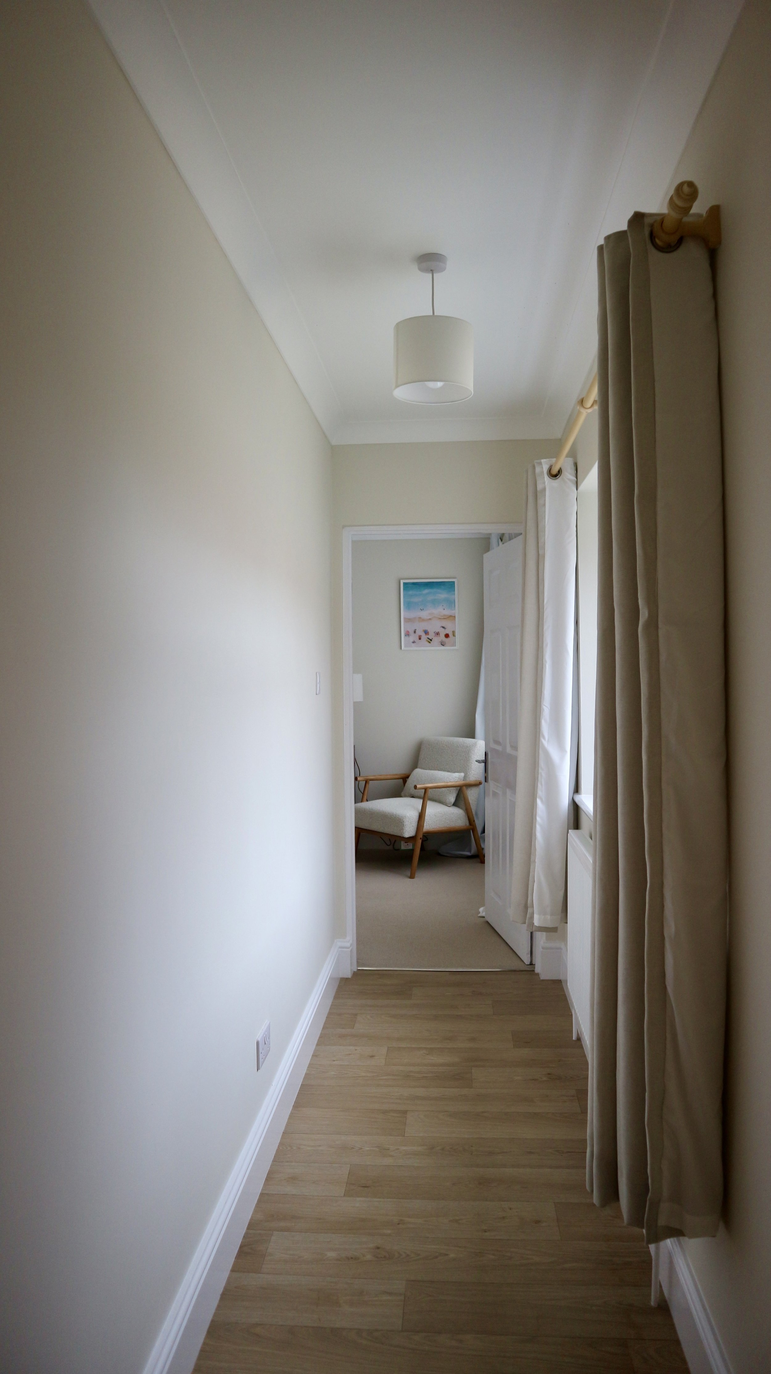 A narrow hallway with light-coloured walls, a wooden floor, a ceiling light fixture, and a window with beige curtains leading to a room with an armchair and a framed picture on the wall. Blackbird Cottage, recently refurbished.