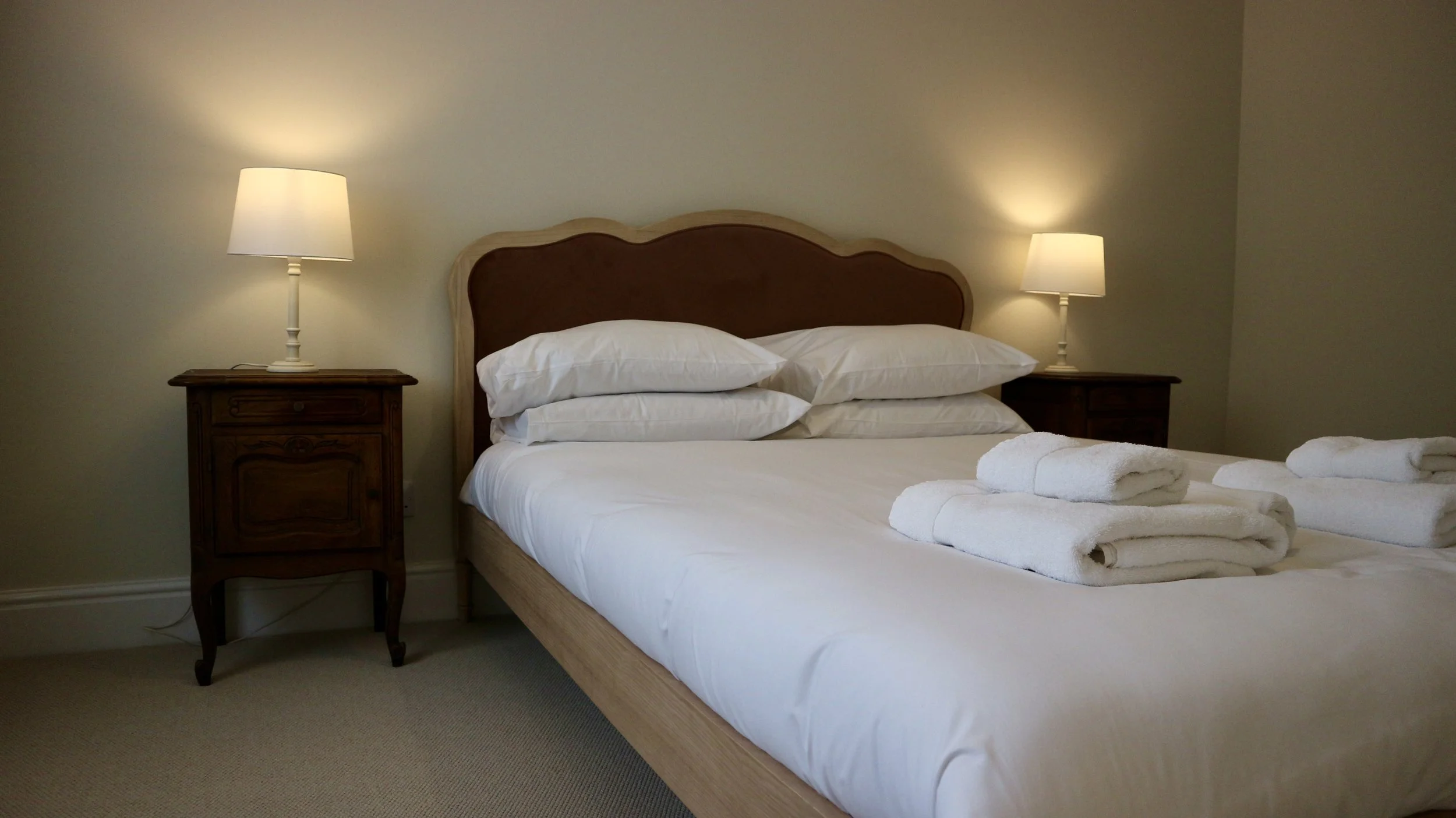 The Master bedroom with white linens and multiple pillows, flanked by two dark wooden nightstands with lamps, in a cosy bedroom. Blackbird Cottage, recently refurbished.