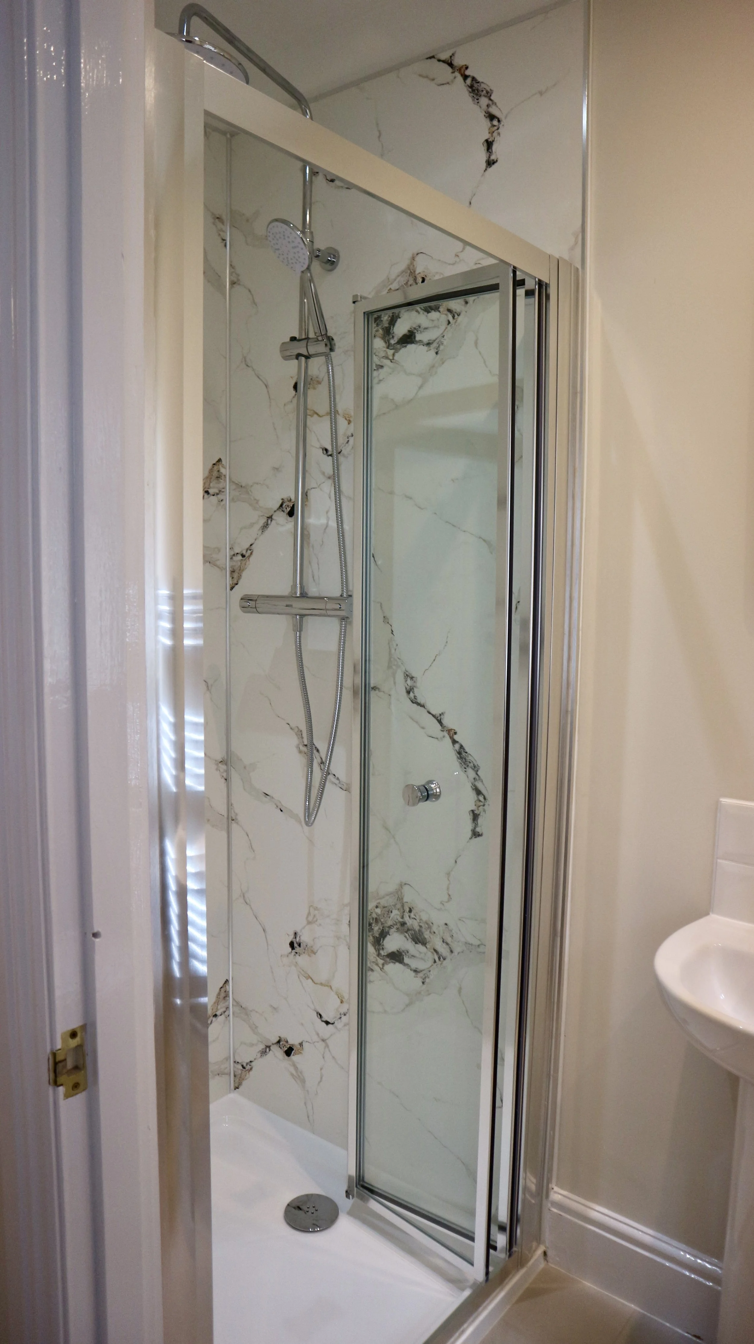 Small shower stall in the master en suite with marble-style patterned tiles, glass door, and handheld showerhead inside, adjacent to a white wall and small sink.