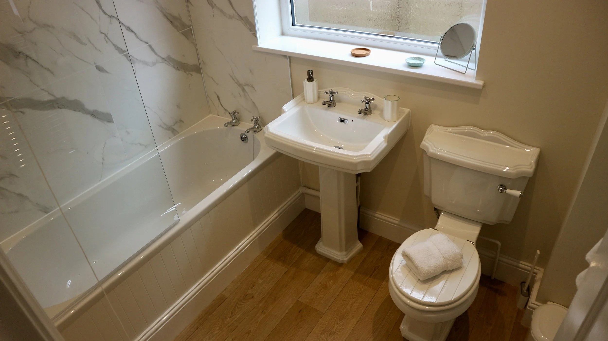 Family Bathroom with bathtub, pedestal sink, toilet, and window, decorated in neutral tones and wooden flooring. Blackbird Cottage, recently refurbished.