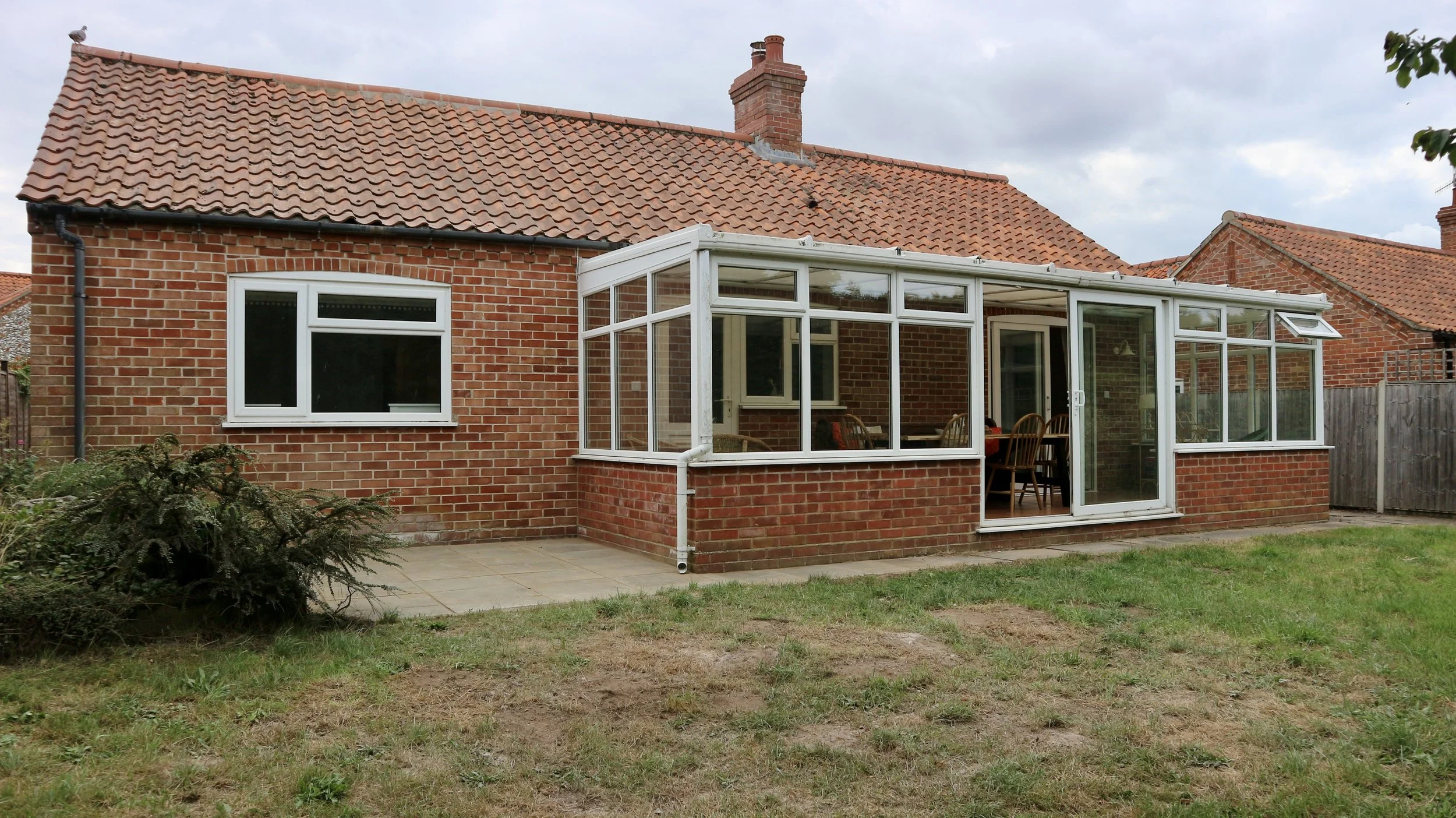 Backyard view of a Blackbird Cottage, recently refurbished. With a tiled roof and a glass-enclosed patio. The yard has grass and a small paved area.