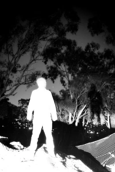 Silhouetted figure in a forest at night, with bright white light creating a stark contrast, surrounded by trees and a dark sky.