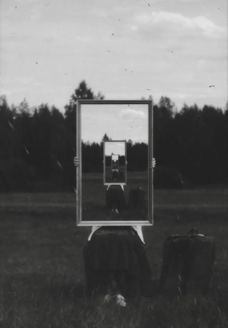 Black and white image of a person holding a large mirror in a grassy field, reflecting the scene infinitely.