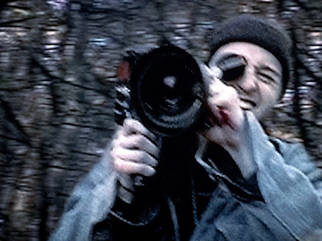 Blurry image of a person looking through a video camera in a wooded area.