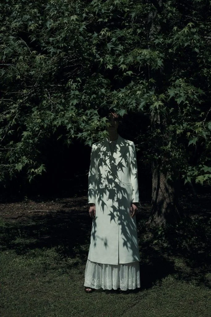 Person wearing a long white coat and lace dress partially obscured by tree leaves in a shaded outdoor setting.
