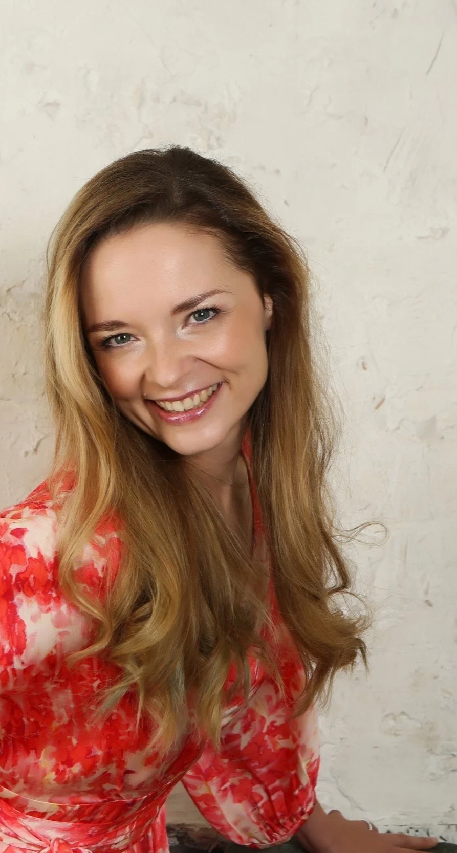 Lächelnde Frau mit langen, blonden Haaren in einem rot gemusterten Kleid vor einer hellen Wand.