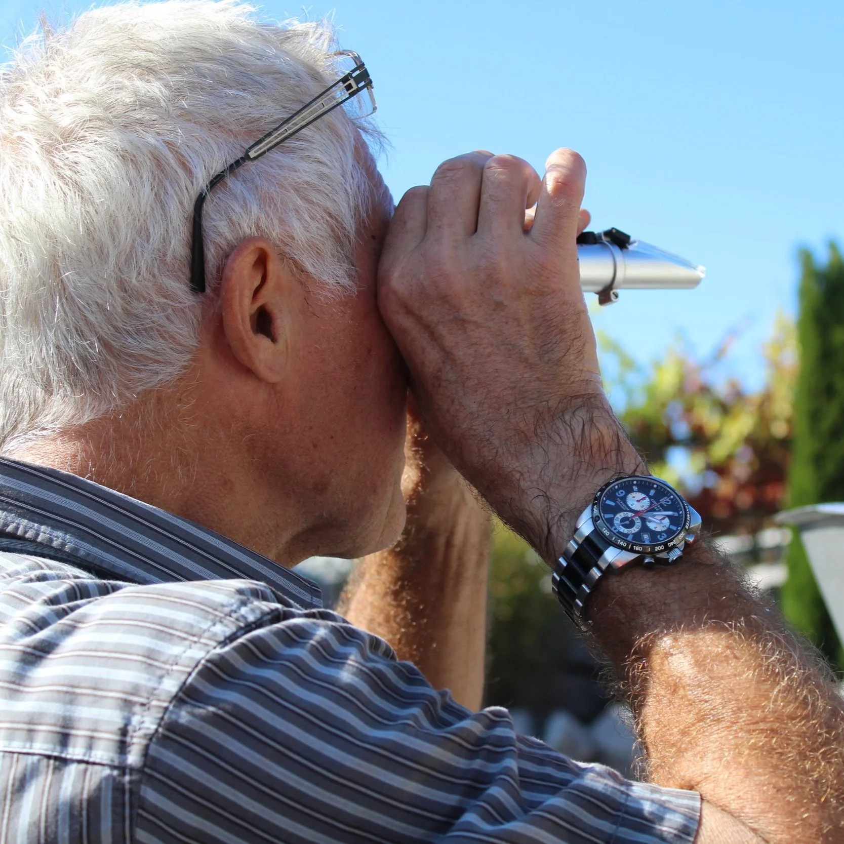 Älterer Mann mit Brille und Uhr schaut durch ein Fernrohr, im Freien bei sonnigem Wetter.