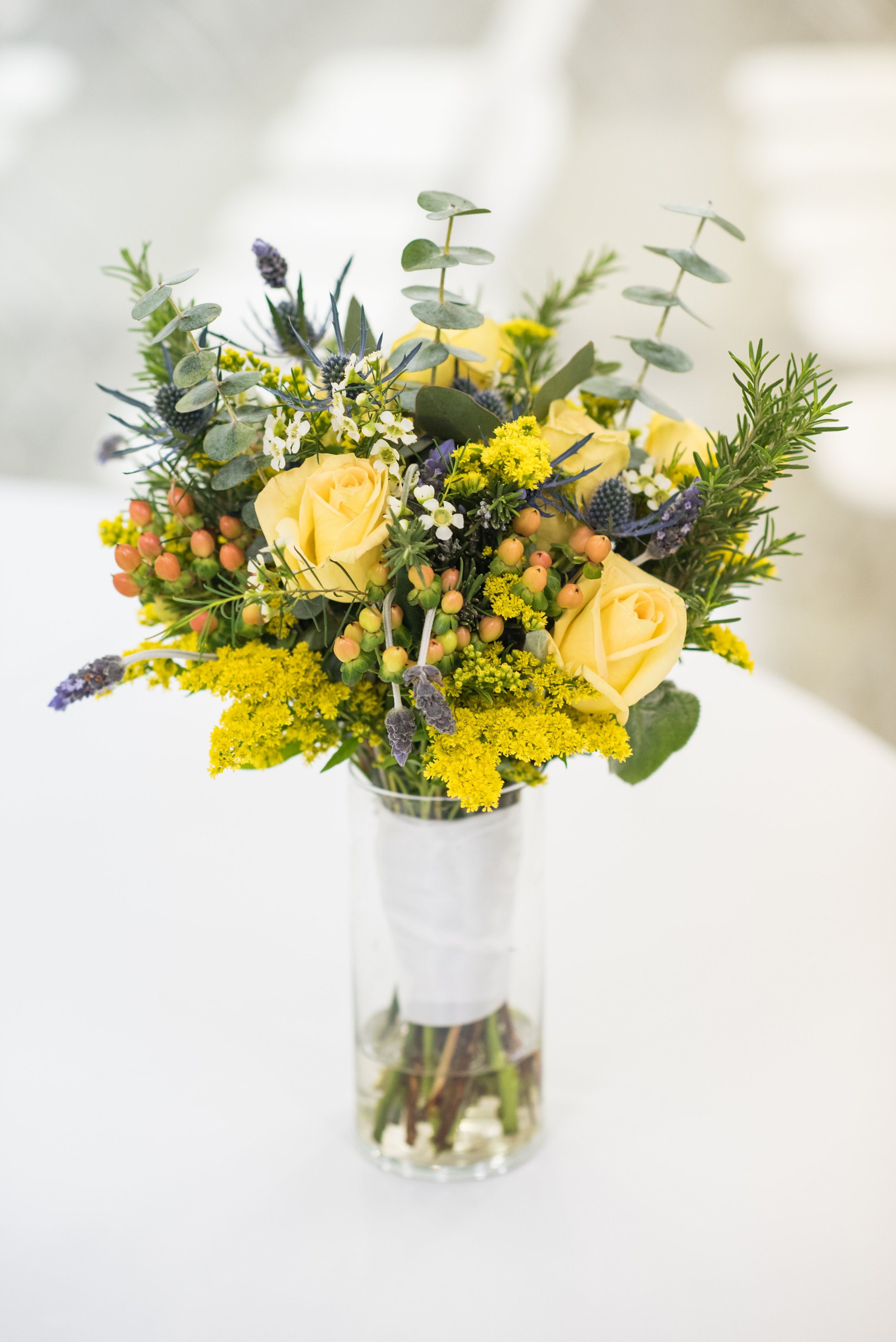 Bouquet of yellow roses with assorted greenery and small colorful flowers in a glass vase on a white table.