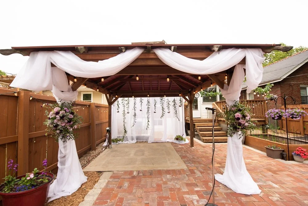 Decorated outdoor wedding pergola with draped white fabric and floral arrangements, surrounded by a wooden fence and brick patio.