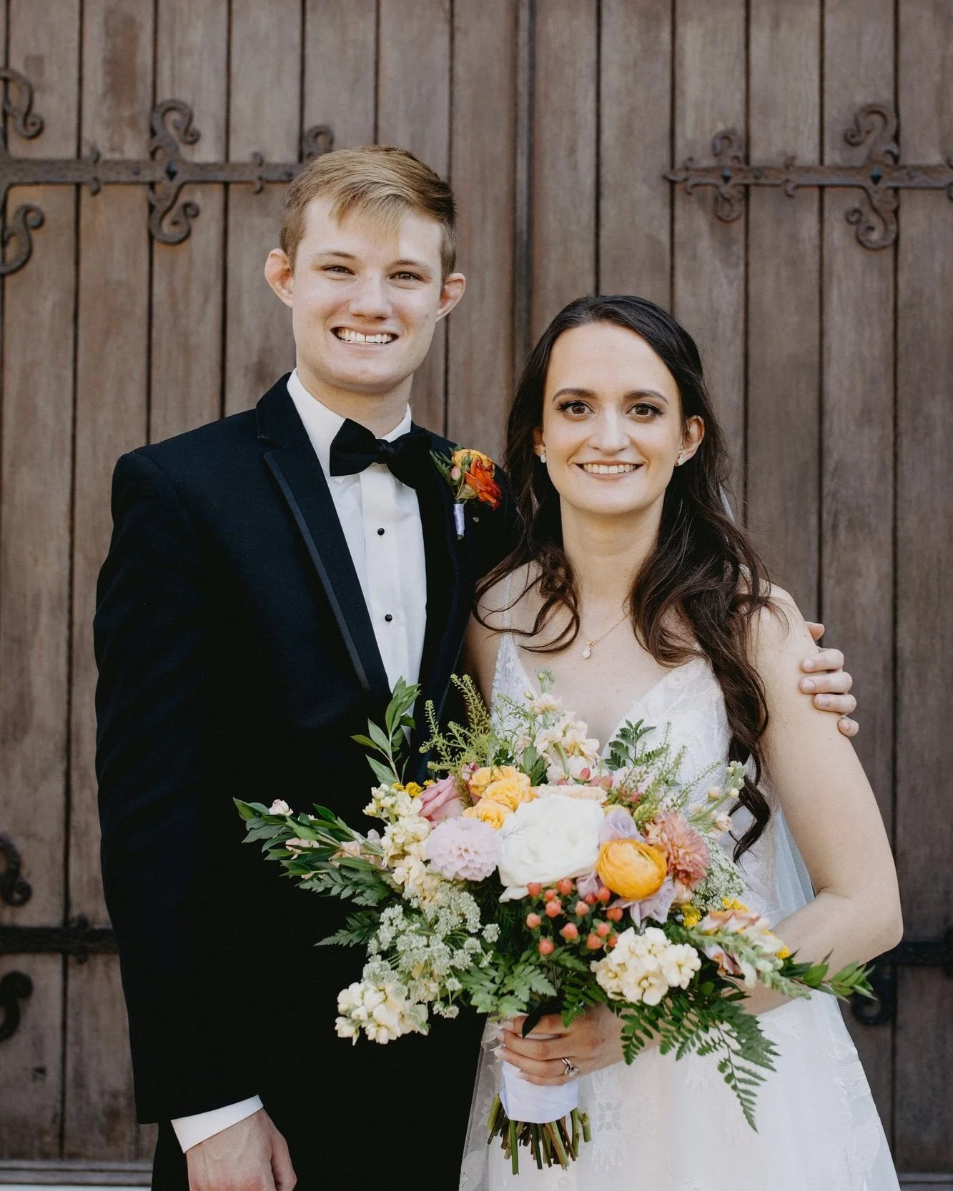 A bride and groom smiling, standing in front of a wooden door. The groom is wearing a black tuxedo with a bow tie, and the bride holds a bouquet of colorful flowers, including roses and peonies, with greenery.