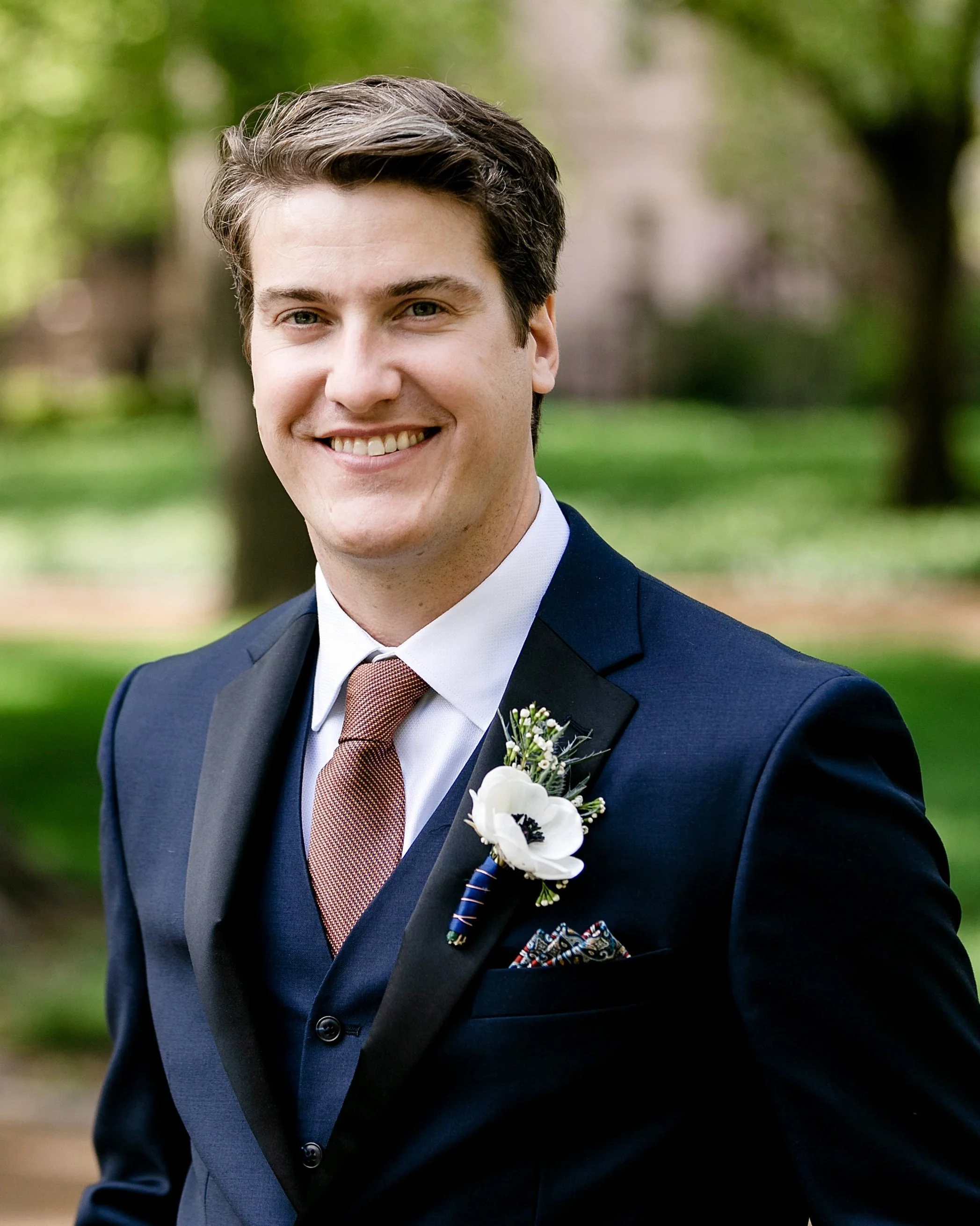 Smiling man in a formal suit with a boutonniere, outdoors.