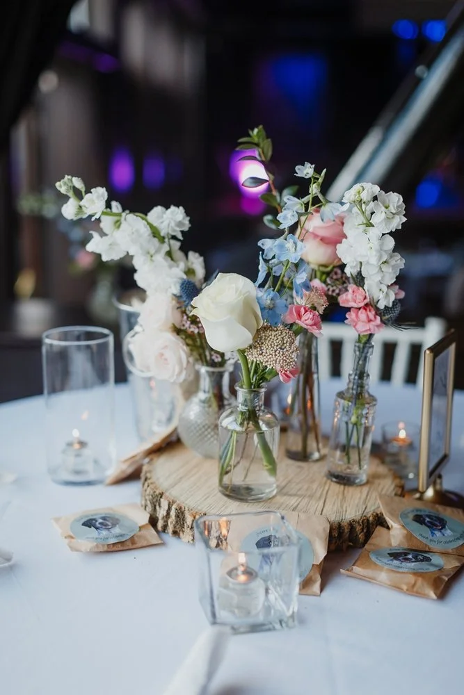 Elegant table centerpiece with assorted flowers in glass bottles on wooden slab, surrounded by candles and small packages on a white tablecloth.