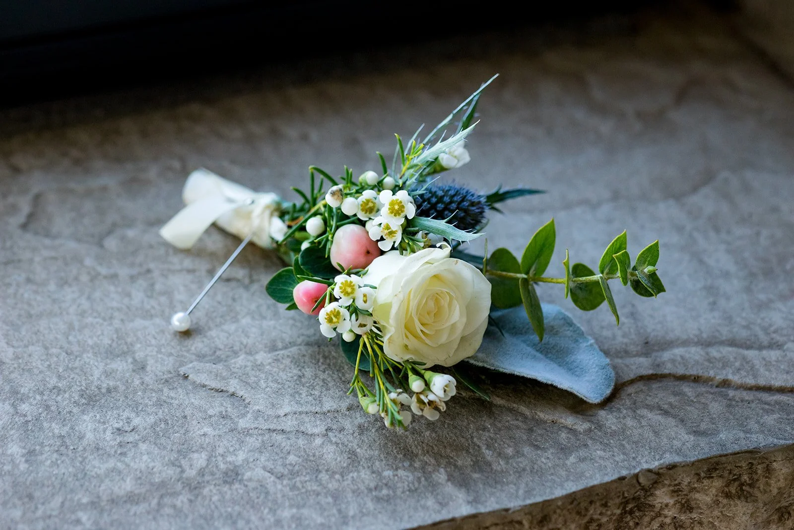 Flower boutonniere with white rose, greenery, and small flowers on stone surface.