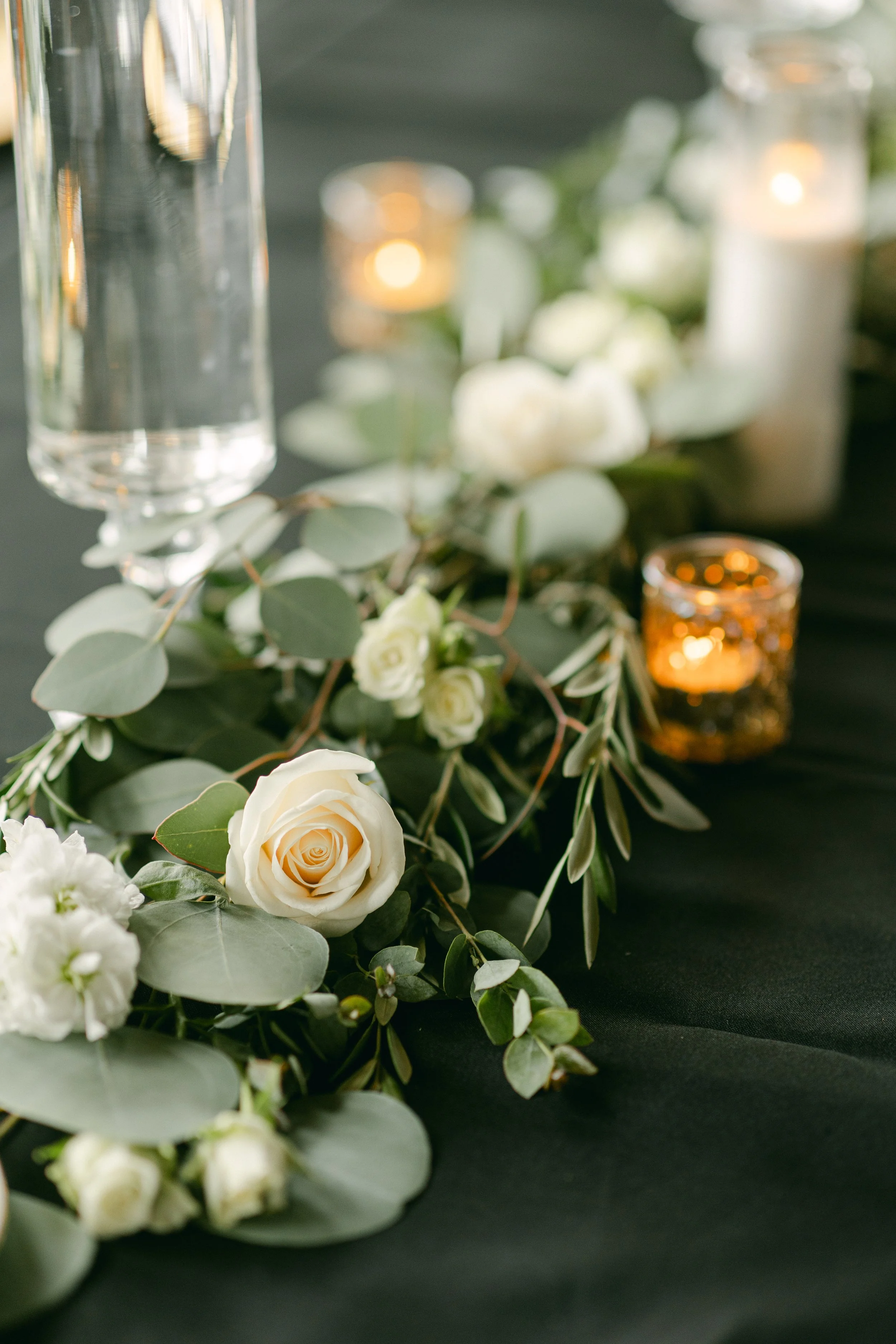 White roses and greenery arranged with candles on a dark tablecloth