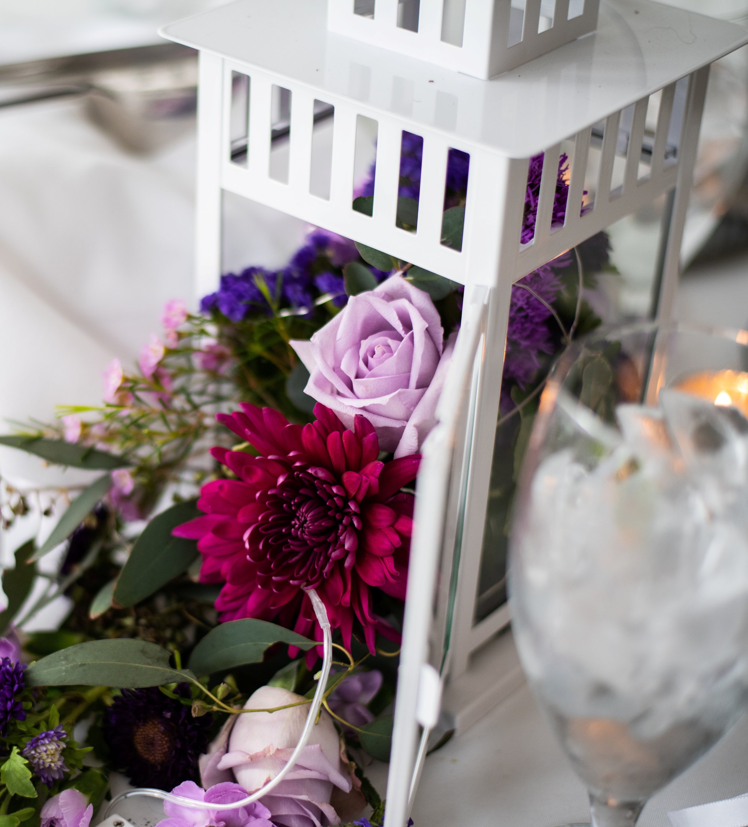 White lantern centerpiece with purple roses, pink flowers, and greenery, alongside a glass of ice water on a table.