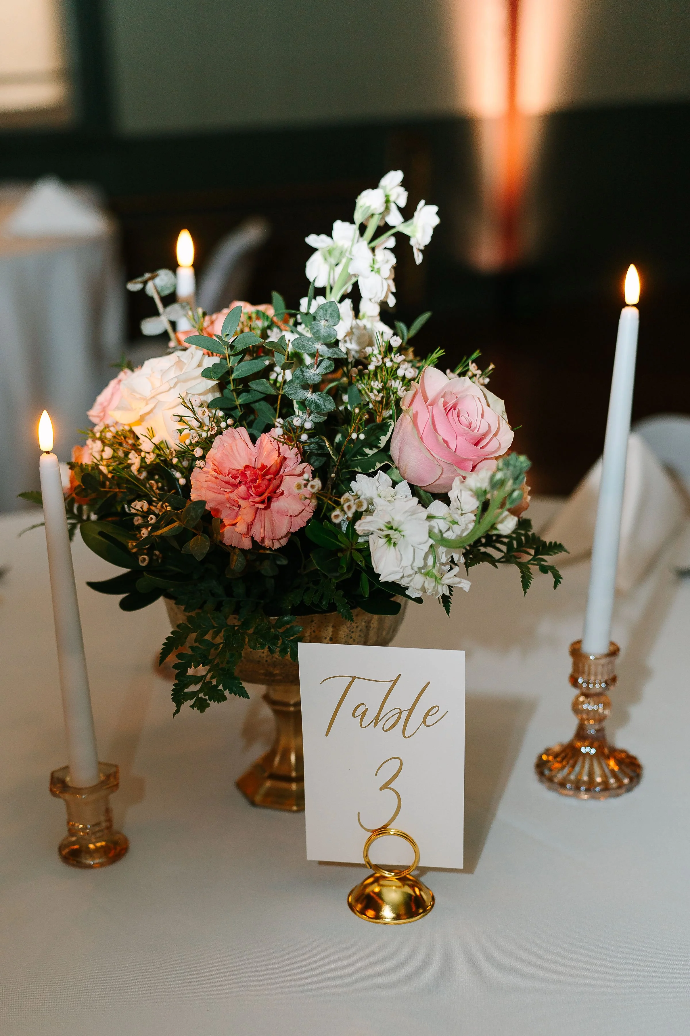 Elegant table setting with floral arrangement, lit tapered candles, and a "Table 3" card on a white tablecloth.