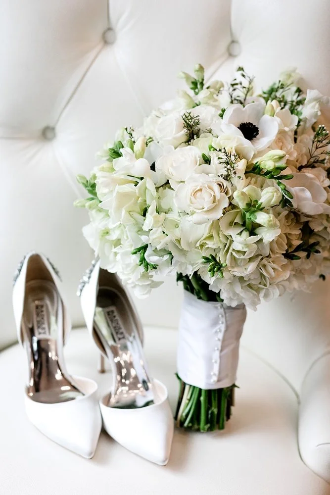White high-heeled shoes and a bridal bouquet of white flowers on a white leather couch.