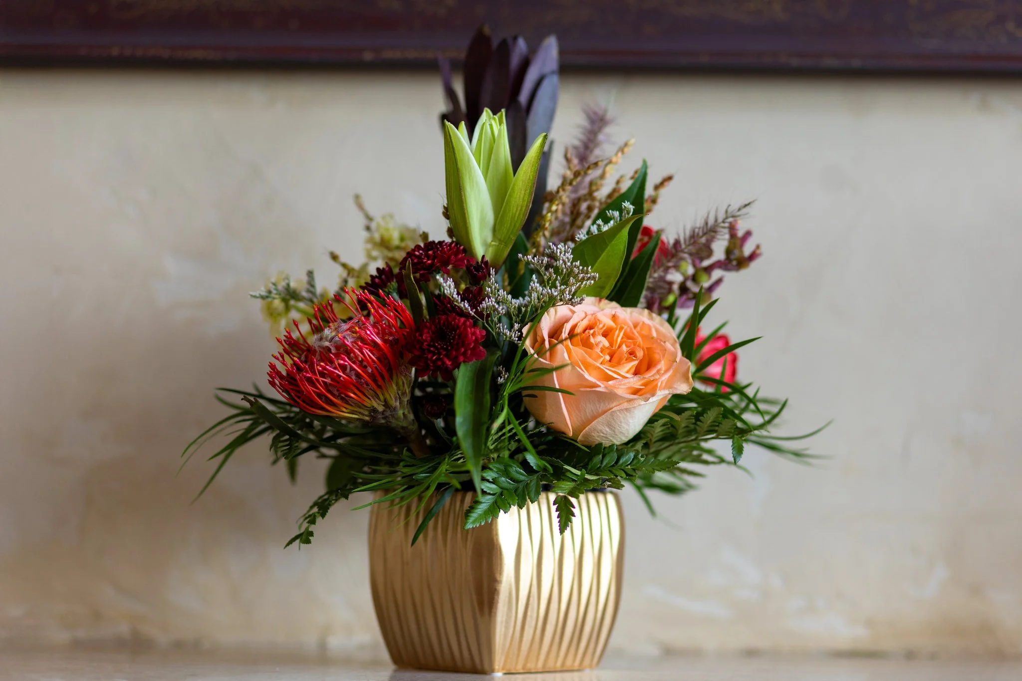 A floral arrangement in a gold vase featuring a peach rose, red pincushion protea, green leaves, and assorted wildflowers against a neutral wall.