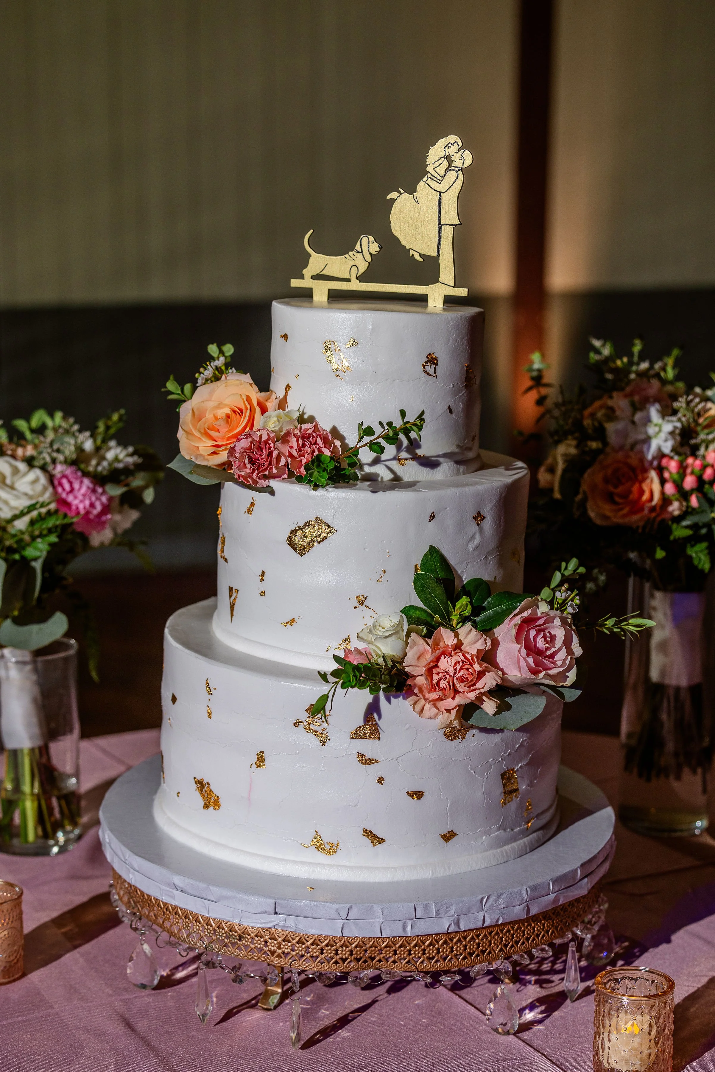 Three-tier white wedding cake with gold accents and floral decorations, topped by a silhouette of a couple and a dog. The cake is displayed on a decorative stand surrounded by floral arrangements.