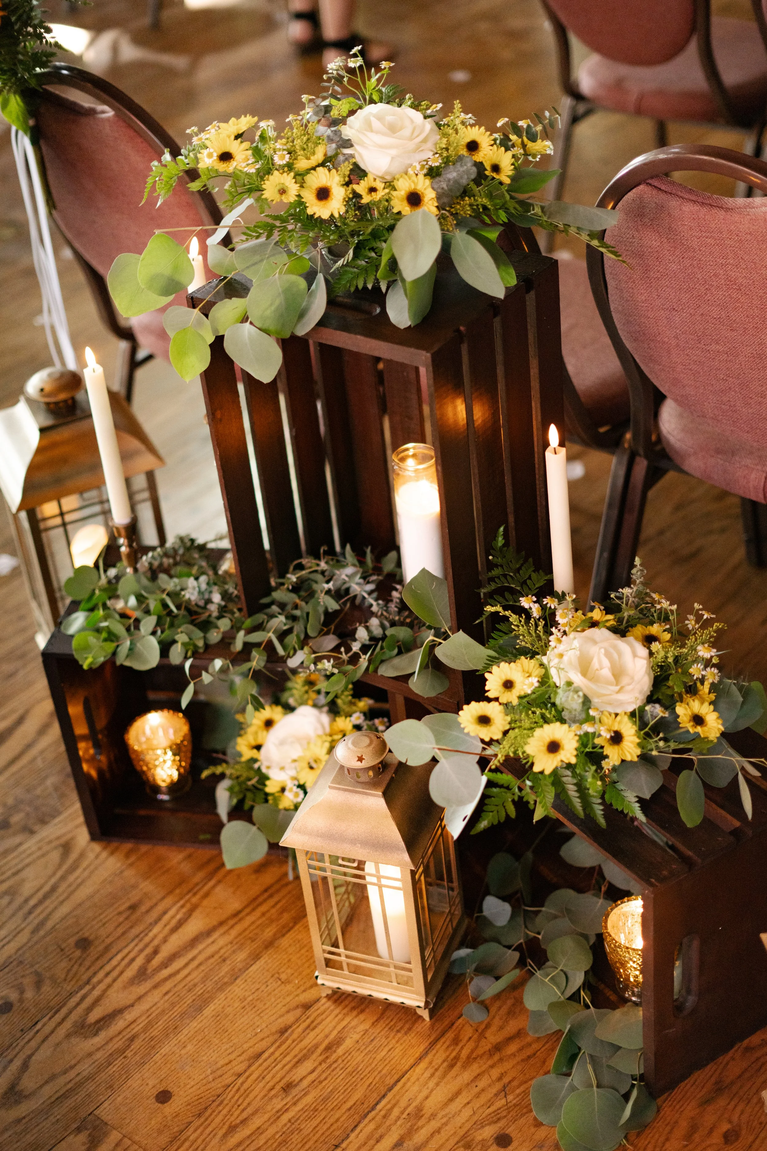 Wooden crates adorned with floral arrangements, including white roses and yellow daisies, surrounded by greenery. Lit candles and brass lanterns are placed nearby, set on a wooden floor.