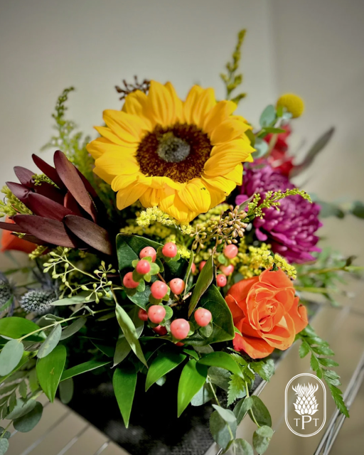 Colorful floral arrangement with a sunflower, roses, berries, and greenery in a black vase.