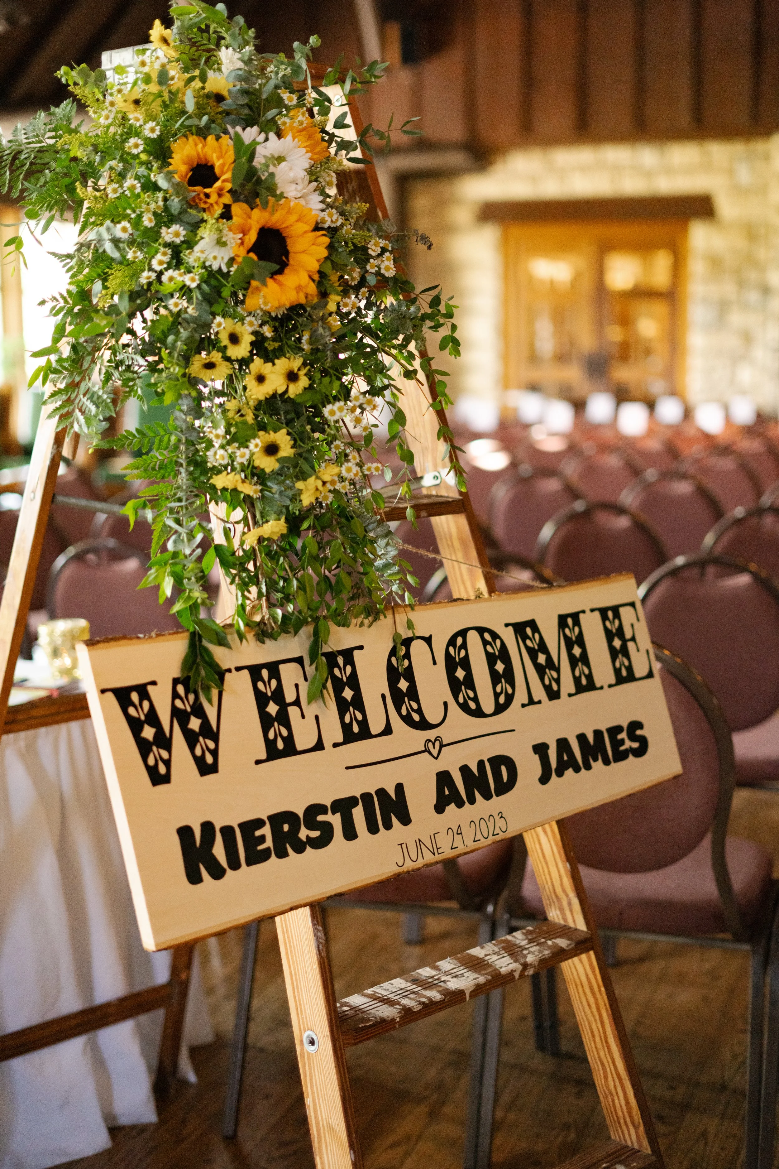 Wooden welcome sign with floral arrangement for a wedding ceremony, featuring sunflowers and greenery, with the names Kierstin and James and date June 24, 2023.
