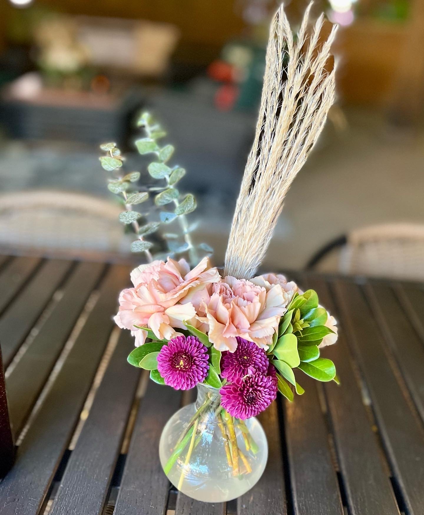 Vase with pink and purple flowers, greenery, and dried grass on a wooden table.
