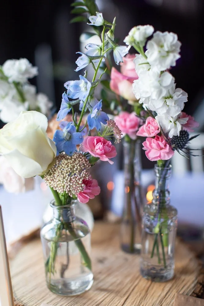 Assorted fresh flowers in glass vases, including blue delphiniums, white roses, pink carnations, and greenery, arranged on a wooden surface.