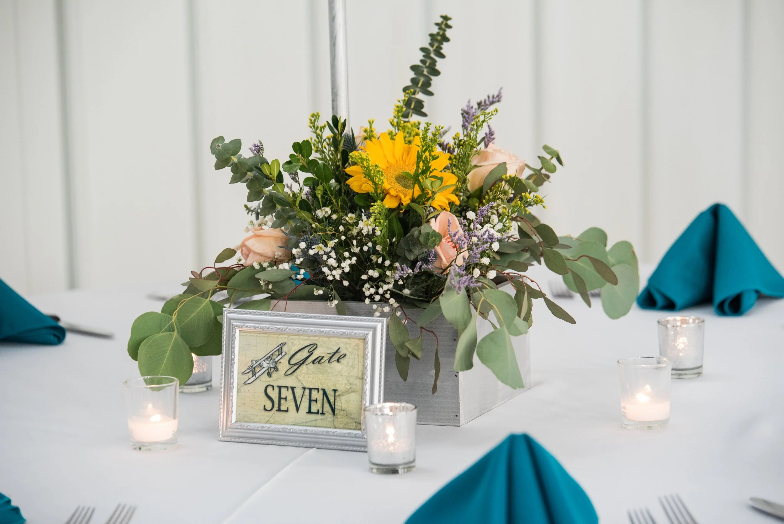 Floral centerpiece with sunflowers and greenery on a table with candles, teal napkins, and a framed table number reading 'Gate Seven.'