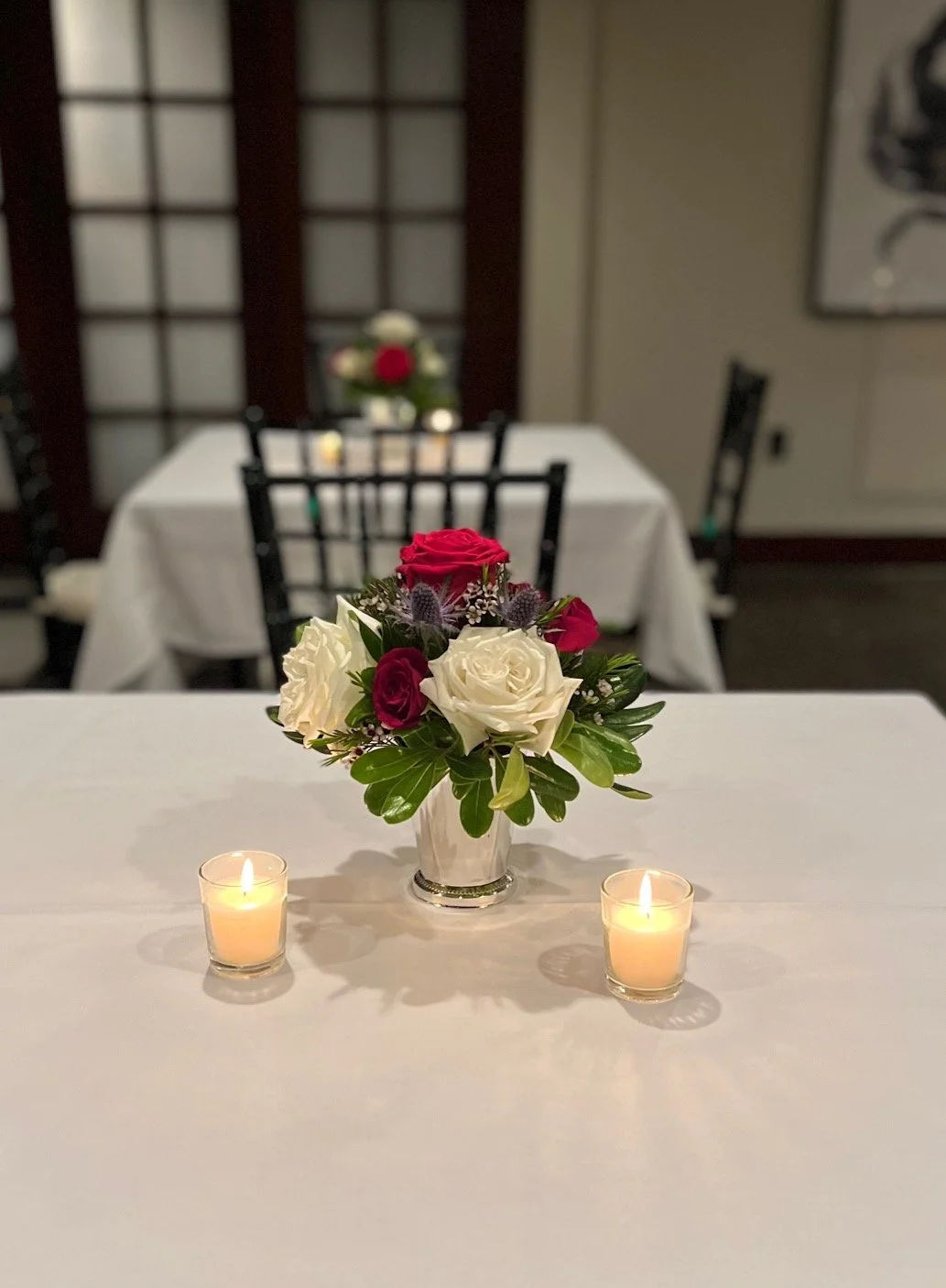 Table setting with a vase of red and white roses and two lit votive candles on a white tablecloth.
