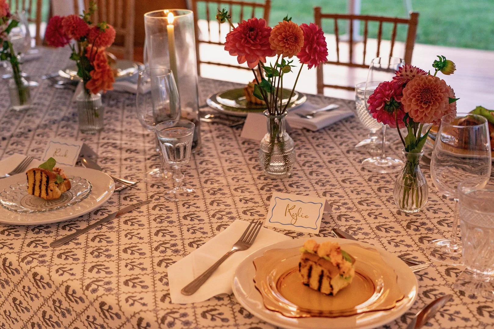 A formal dining table set with pink and orange flowers in small glass vases, candles, glassware, plates with cake, and place cards, in a well-lit outdoor setting.