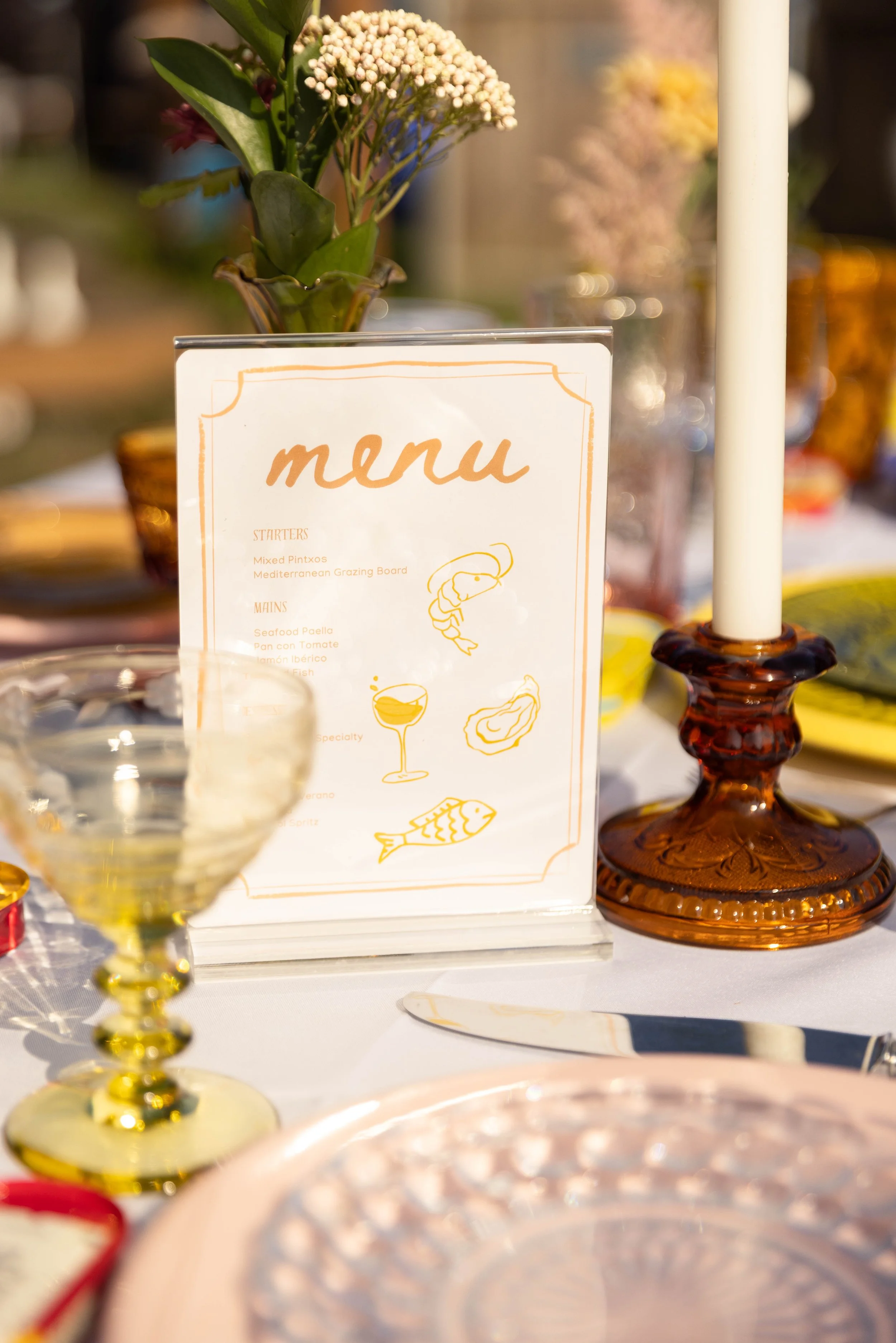 A table setting with a menu card, a lit candle in a glass holder, floral arrangements, a wine glass, and a plate with a decorative pattern, all in warm tones.