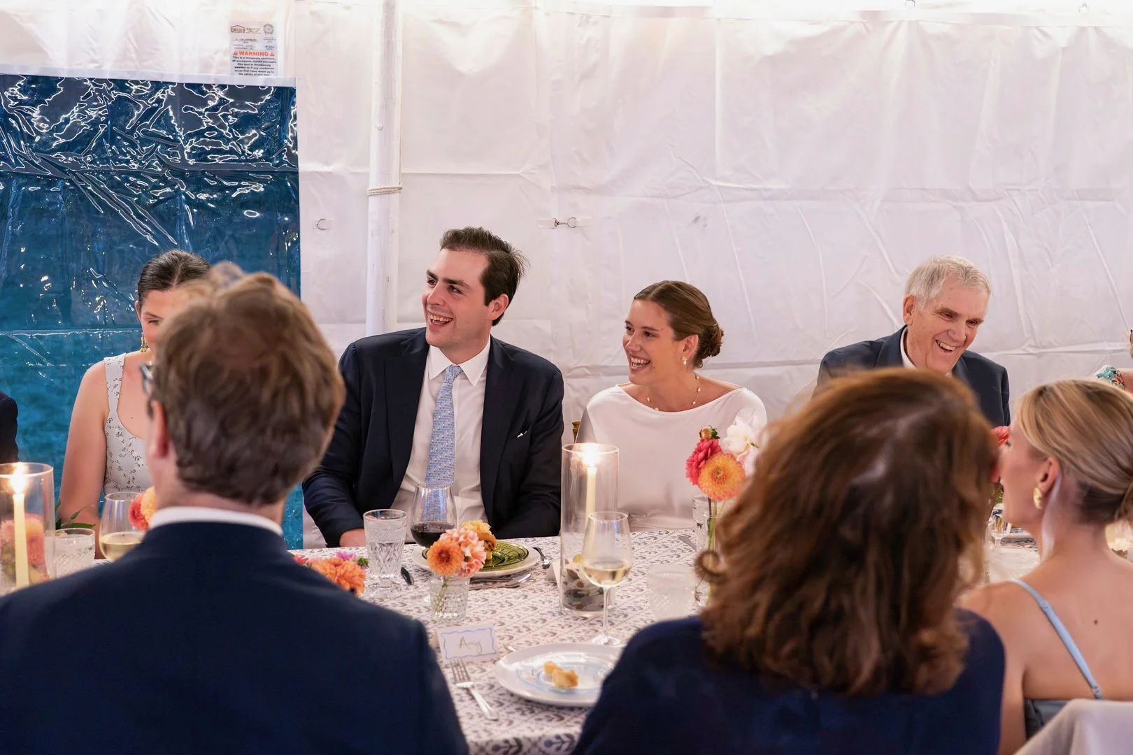 People smiling and talking at a wedding reception table with floral centerpieces, candles, and wine glasses in a tented area.