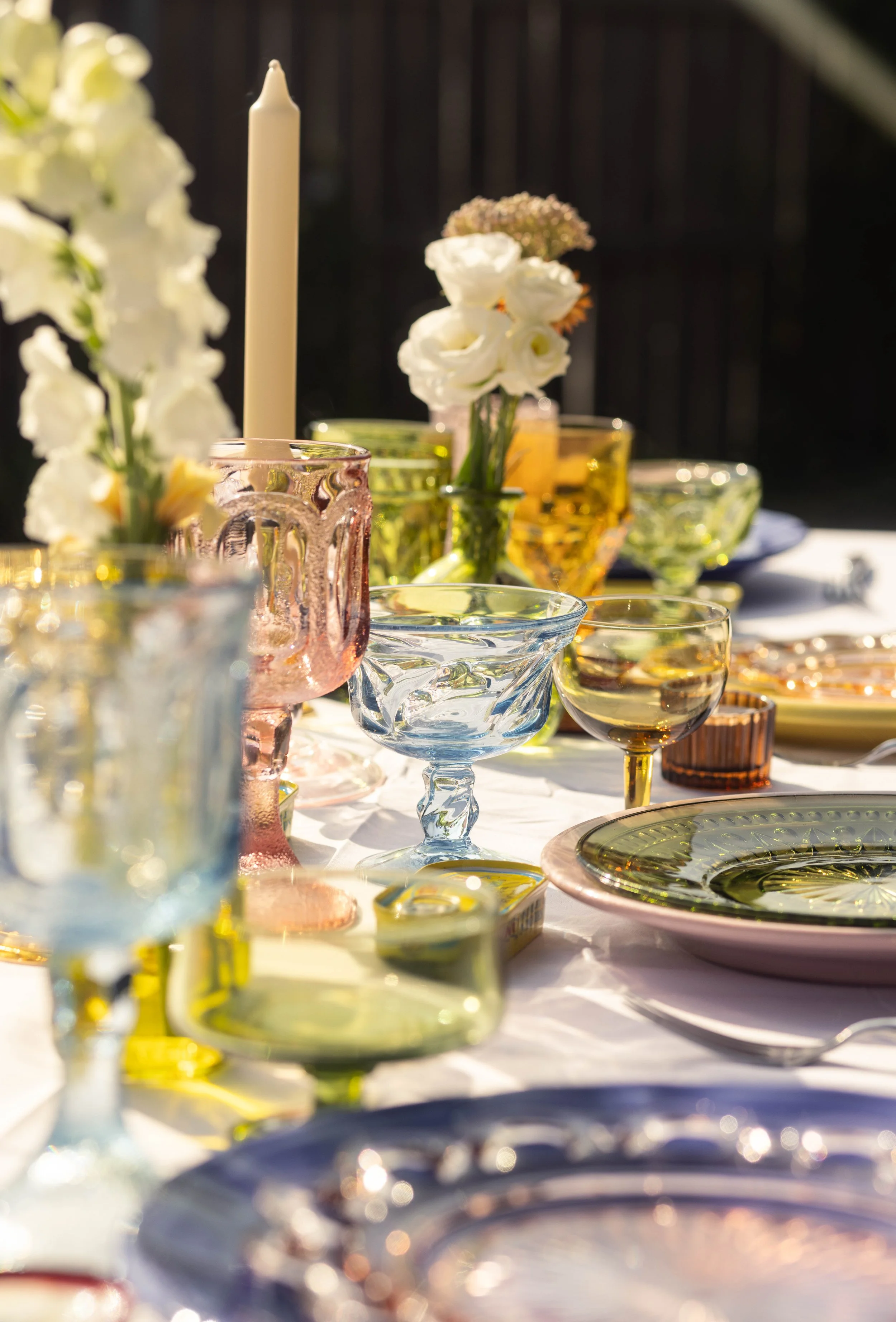 Colorful glassware and floral centerpieces on a white tablecloth, set outdoors with sunlight.