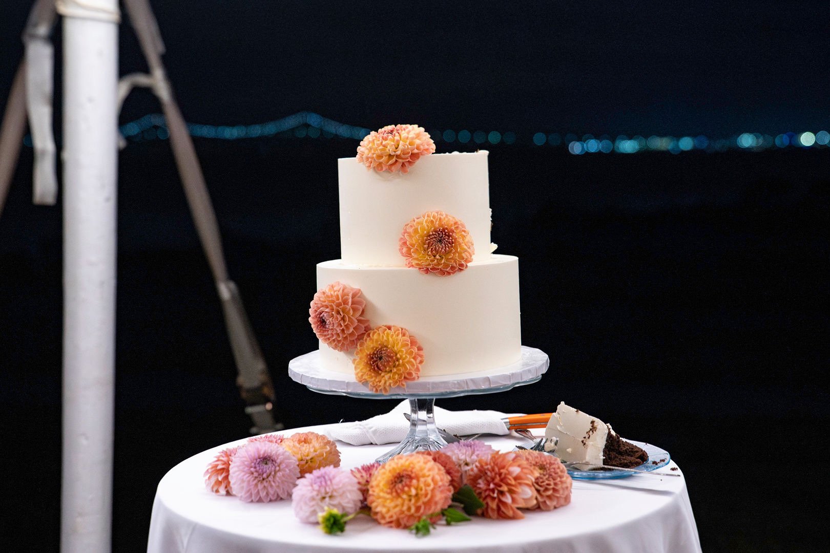 A two-tiered white wedding cake decorated with pink and orange flowers on a glass stand, surrounded by additional flowers on a round table with a slice of cake and a fork against a nighttime background.