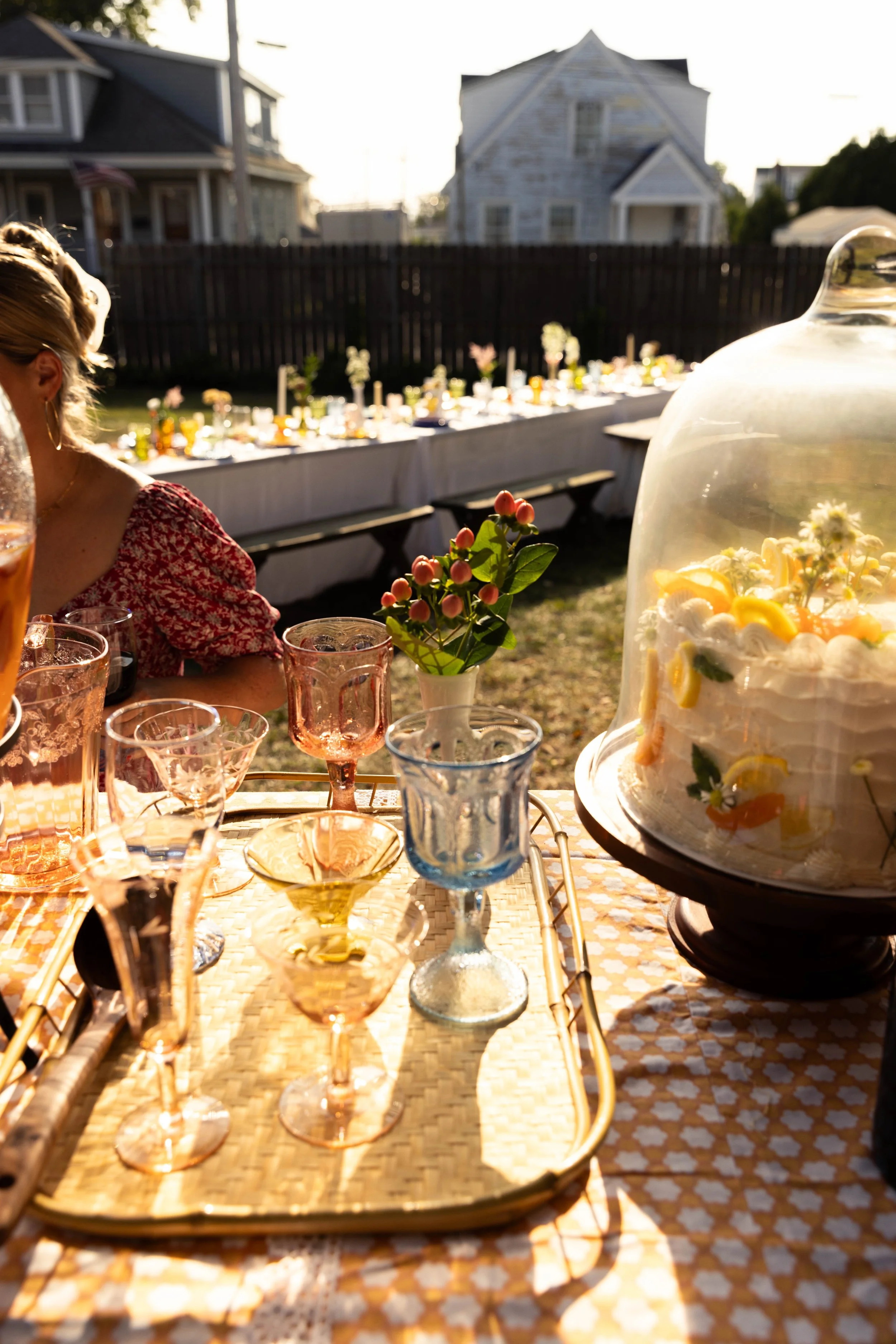 A festive outdoor gathering with colorful glassware, a cake under a glass dome, and a long table decorated with flowers in a backyard during sunset.
