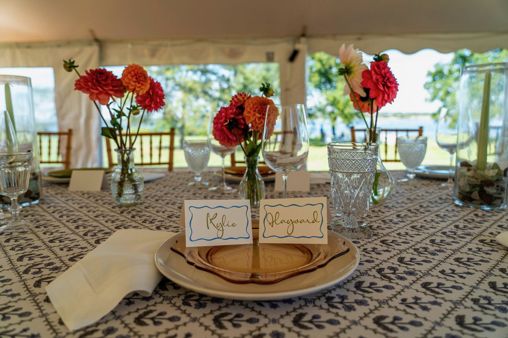 Elegant table setting with pink and orange flowers in glass vases, plates, glasses, name cards, and a patterned tablecloth, set outdoors with trees and water visible in the background.