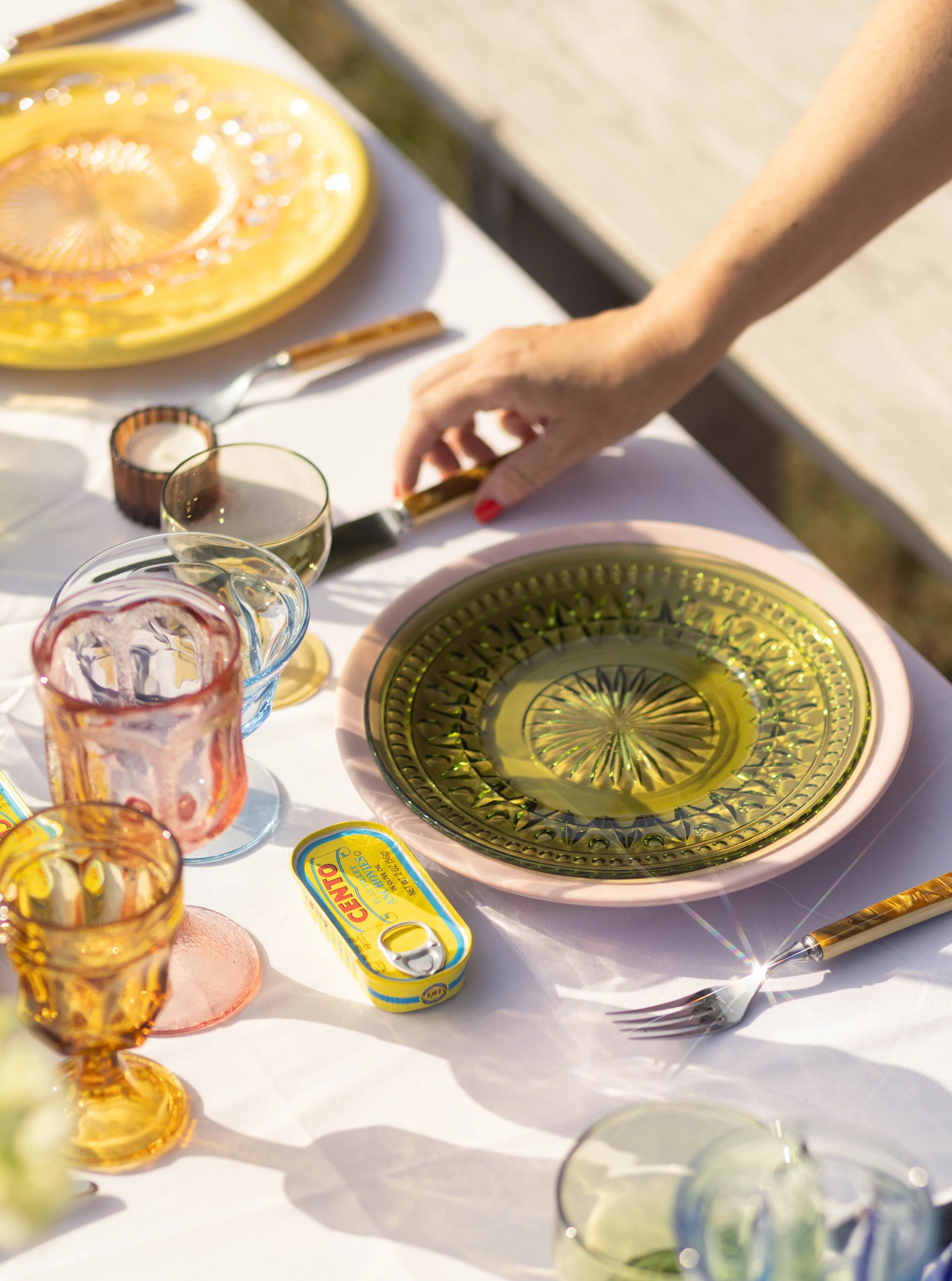 A table set for a meal with colorful glassware, yellow and green plates, and a tin of anchovy fish in the center. A person's hand is reaching for a fork on the table.