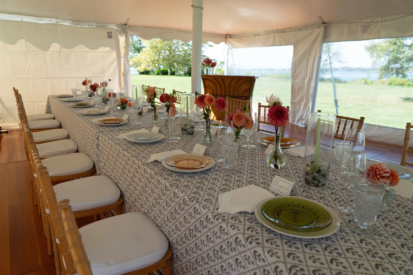 Long banquet table set with pastel-colored plates, glassware, and pink and orange flowers in vases inside a white tent with open sides overlooking a grassy landscape.