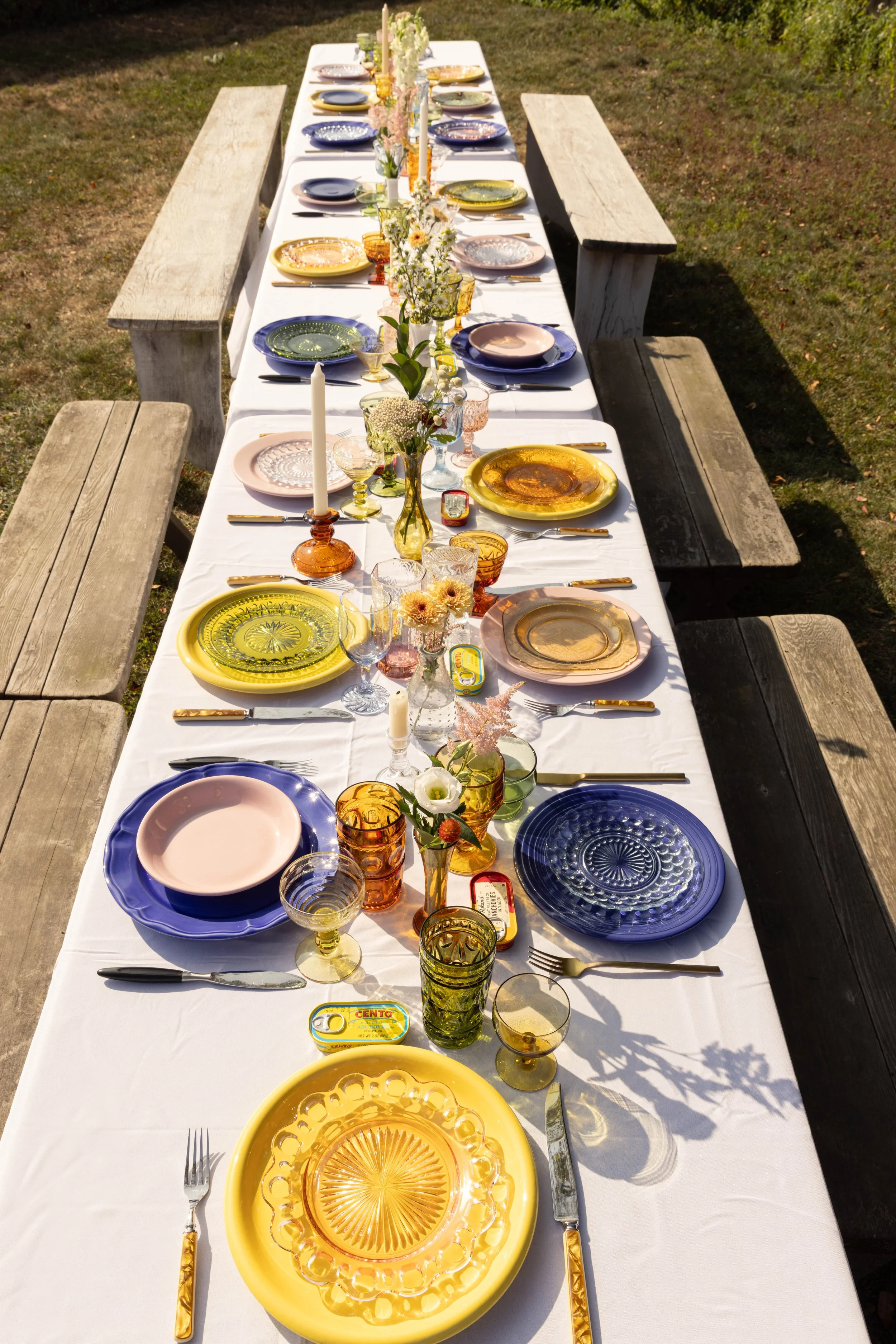 A long outdoor table set for a meal with colorful glassware, plates, flowers, and candles on a white tablecloth, surrounded by wooden benches.