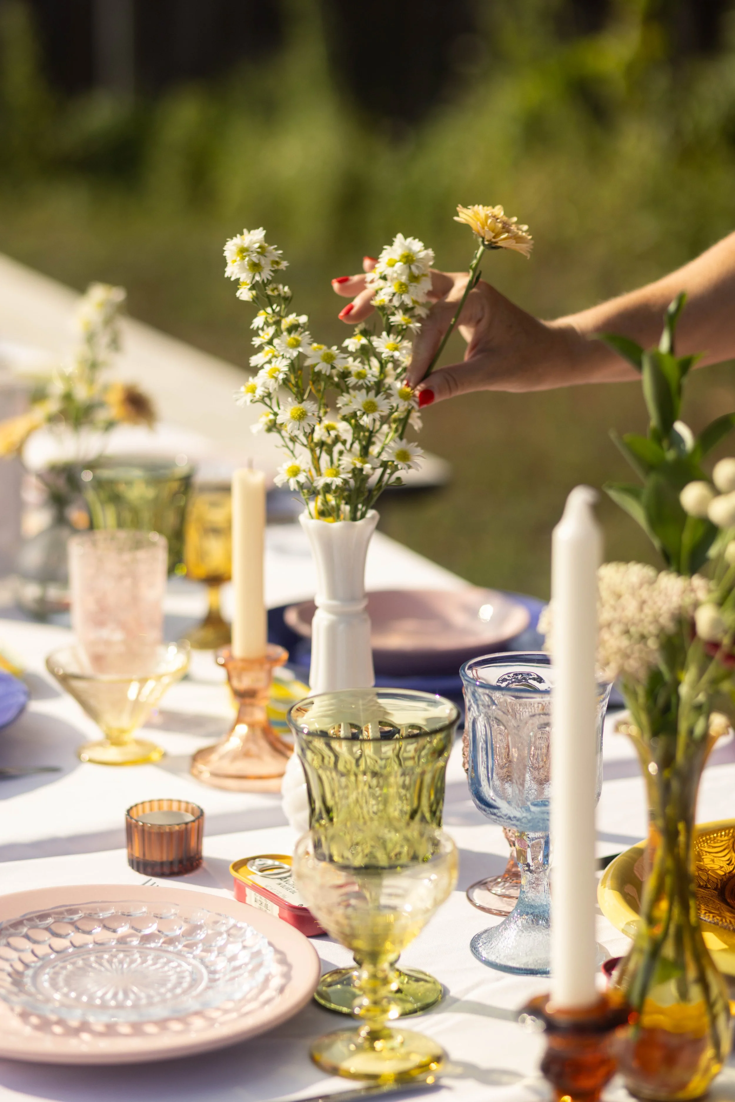 A table set outdoors with colorful glassware, candles, and a white vase holding daisies and other flowers. A person's hand is arranging the daisies in the vase.